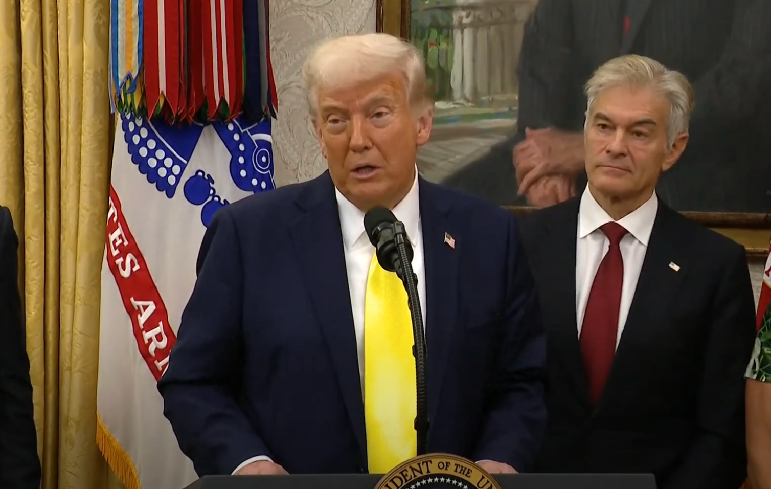 A man in a suit speaks at a podium with a seal, accompanied by another man in a suit, in a formal setting