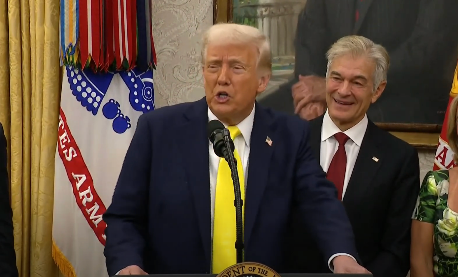 A public figure speaks at a podium in a formal setting, accompanied by others in suits, in front of military-themed decor