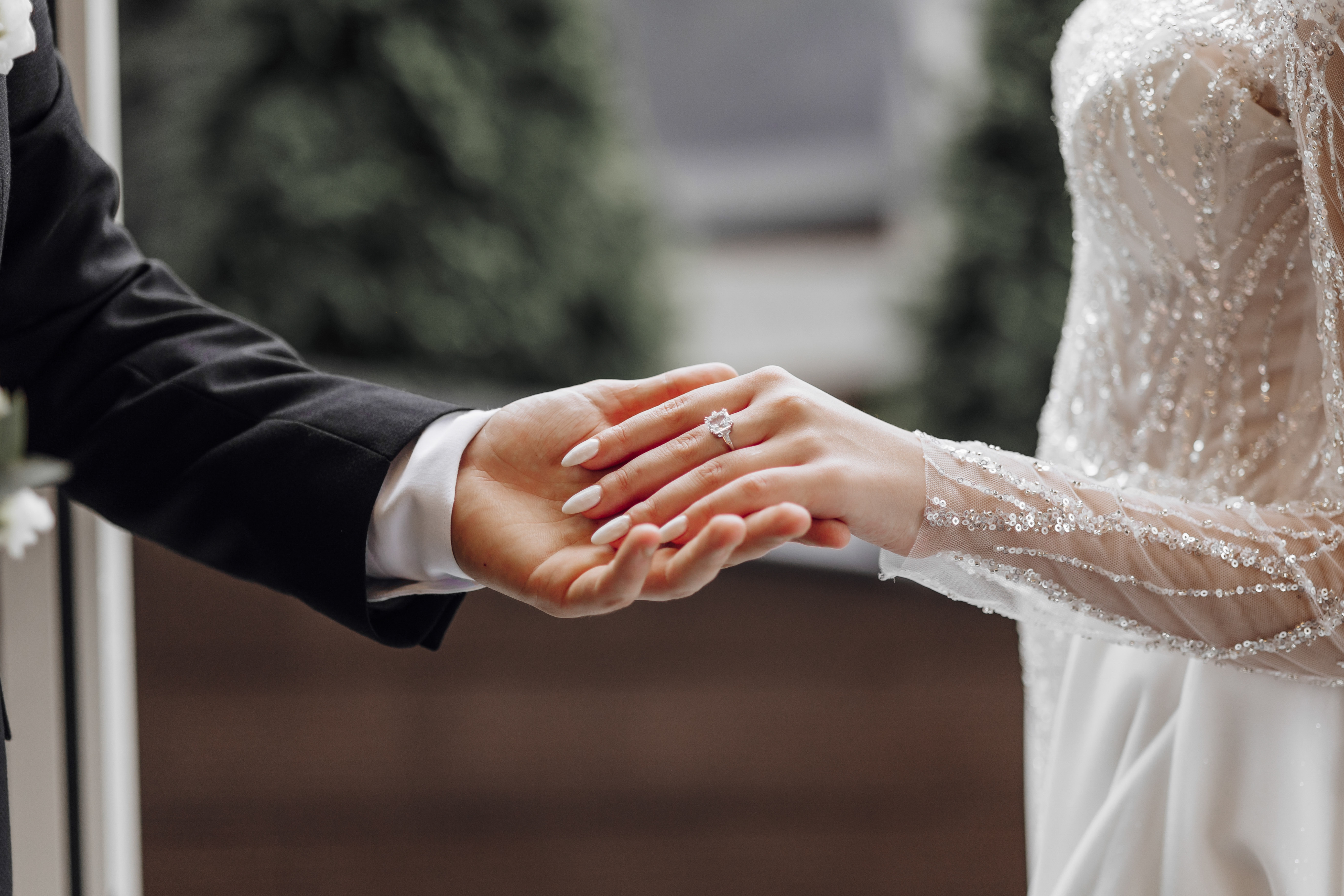A bride and groom holding hands; the bride wears an elegant lace wedding gown and an engagement ring