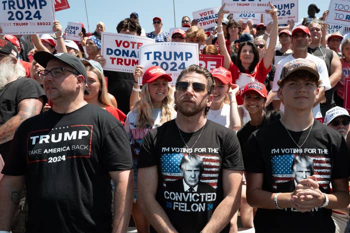 A crowd at a rally, many wearing pro-Trump shirts and hats, holding signs supporting Donald Trump for the 2024 presidential election