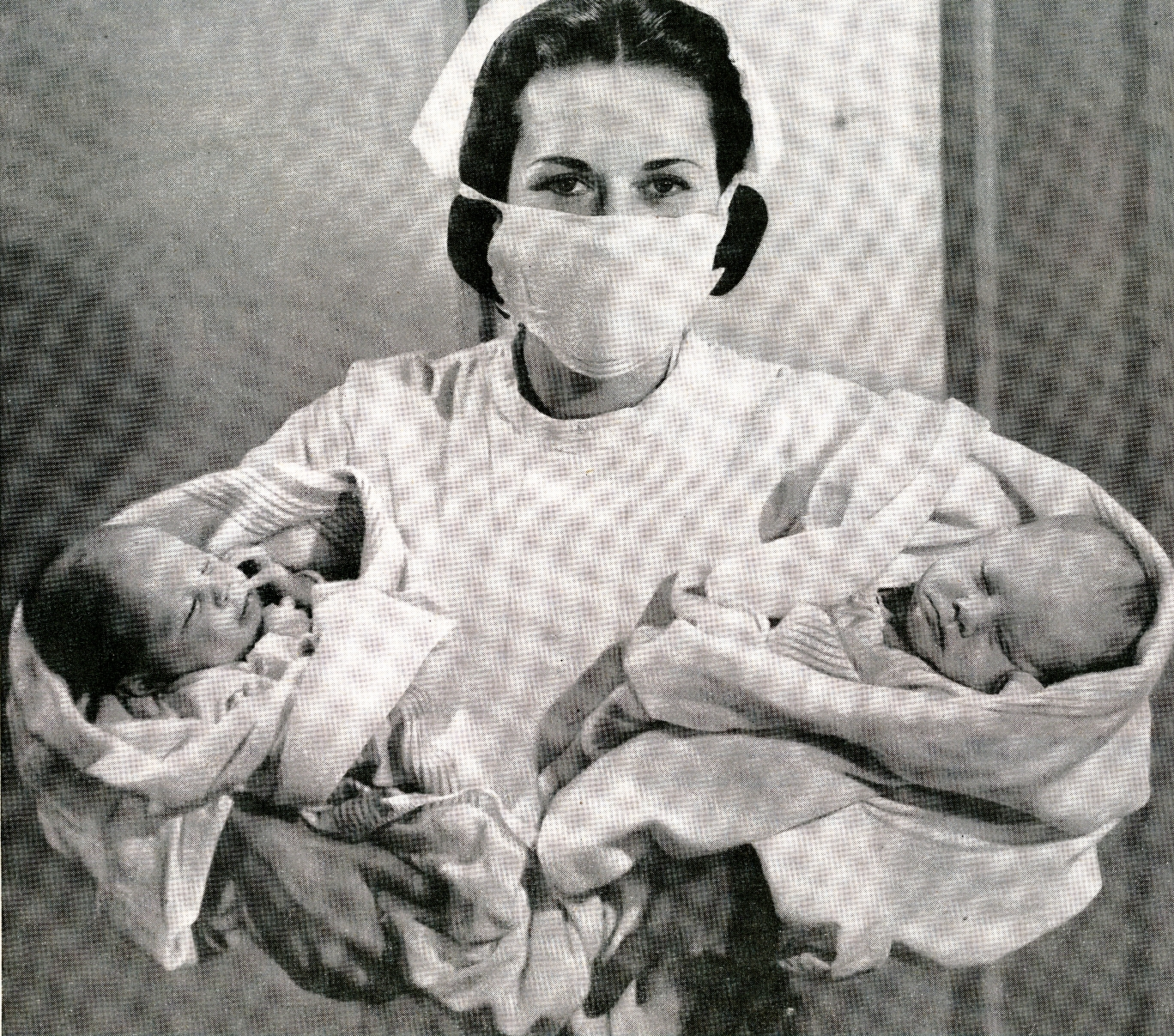 Nurse holds two newborn babies, each wrapped in blankets, in a hospital setting
