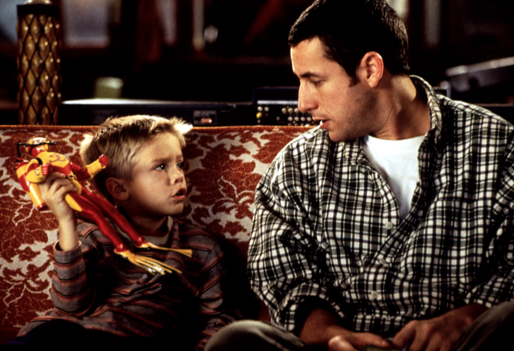 Man in a plaid shirt talks to a young boy on a sofa. The boy holds an action figure, looking attentive. Indoor setting, casual interaction