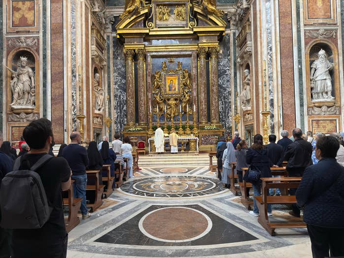 People attending a religious service in an ornate church with statues and a gold altar