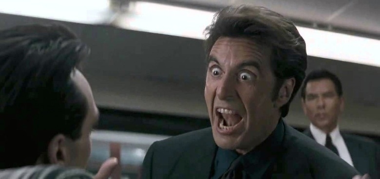 A man with an intense expression is speaking emphatically in a busy indoor setting. Another person is visible in the background