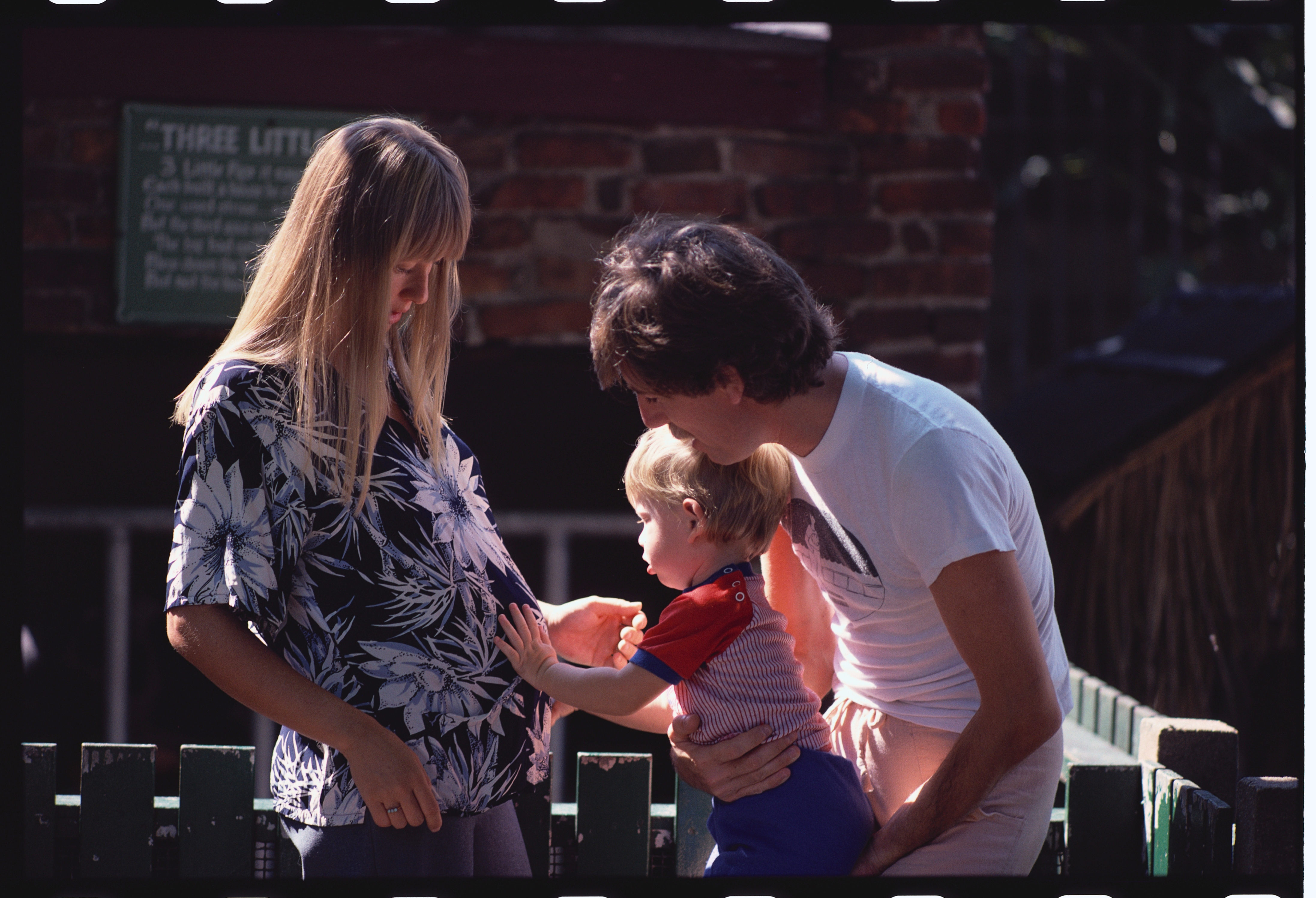 A man holds a young child who is touching a pregnant woman's belly, creating a tender family moment outdoors