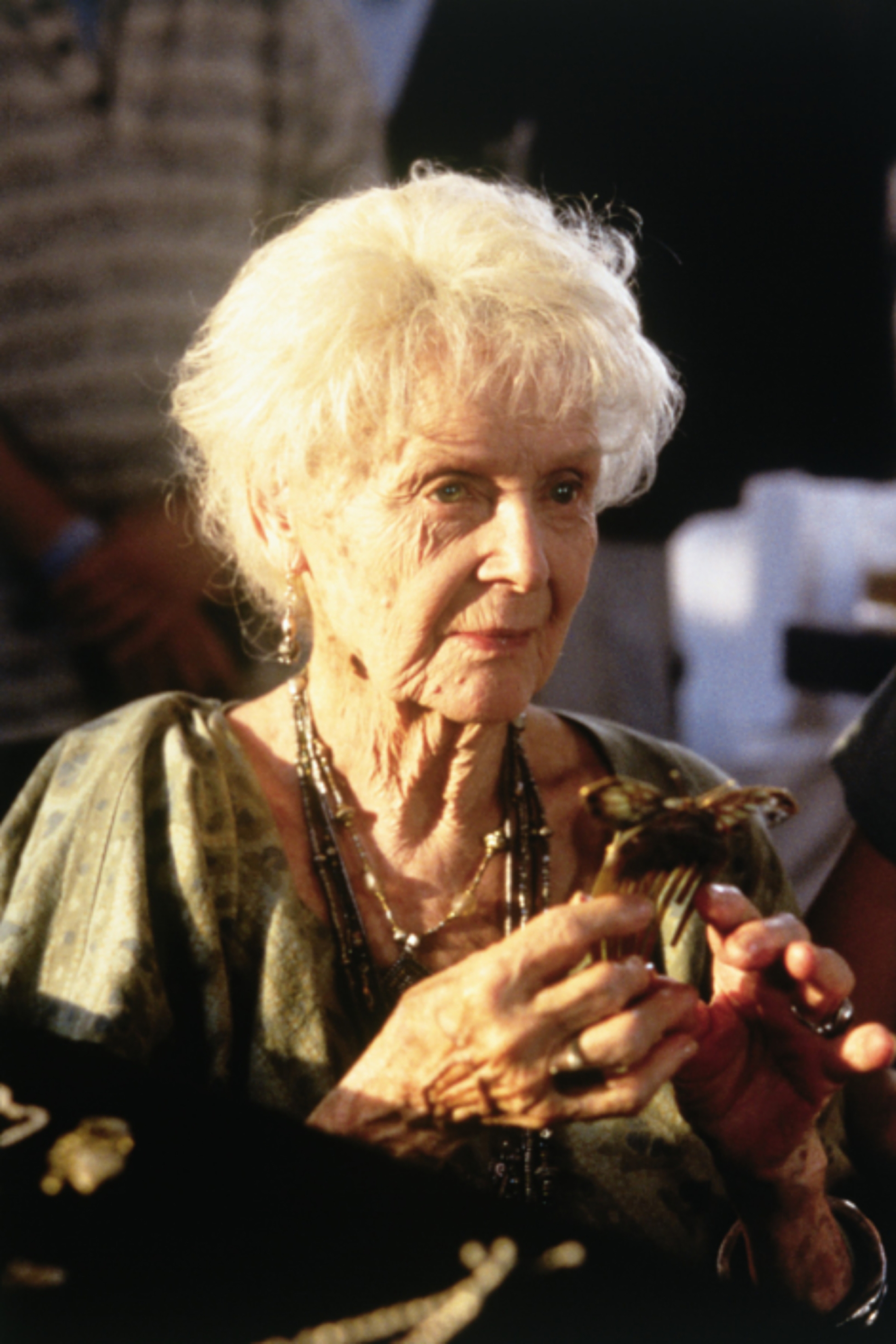 An elderly person with short, white hair holds an object, wearing a patterned shawl and layered necklaces