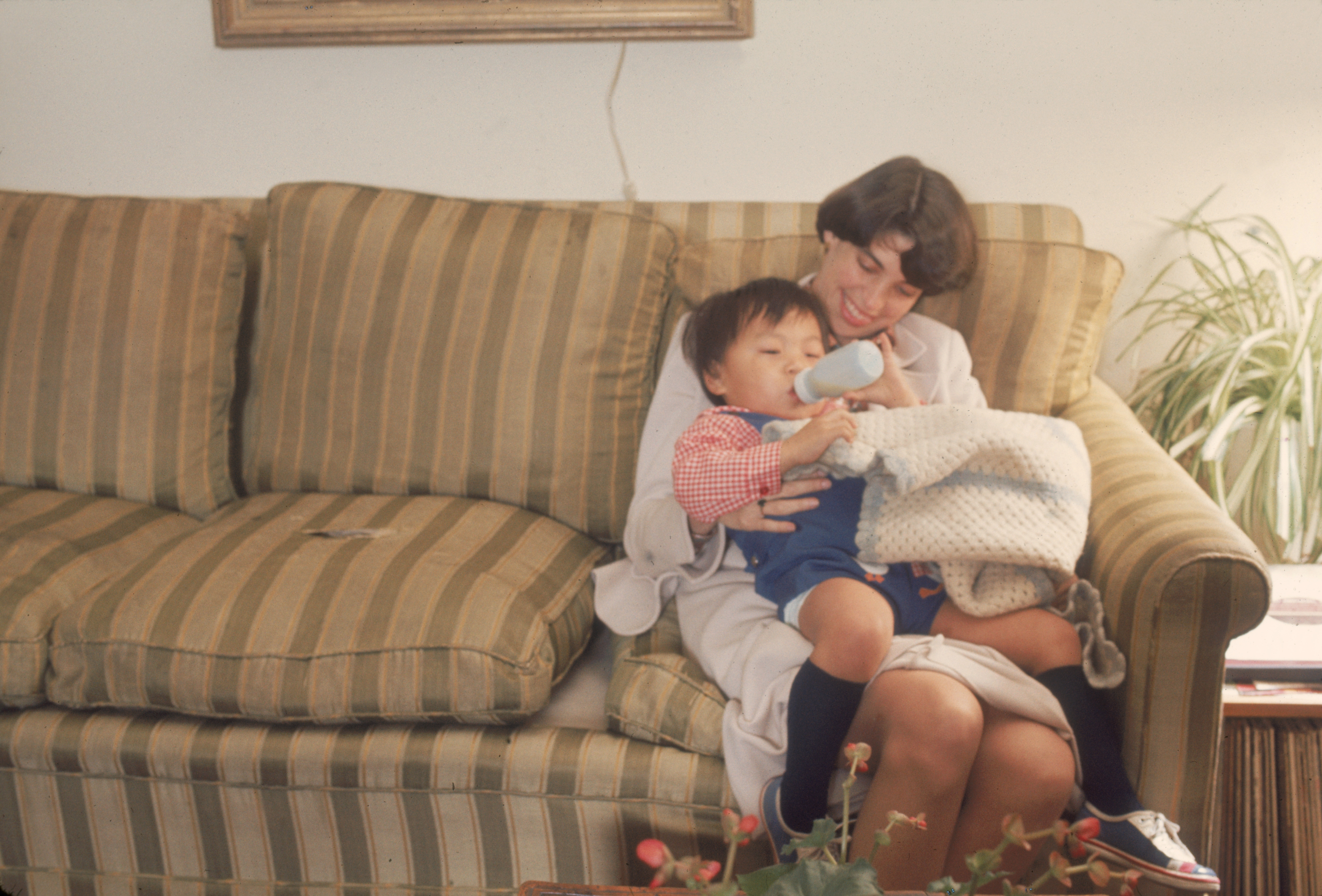 Woman on a couch bottle-feeding a young child wrapped in a blanket. Cozy and nurturing scene