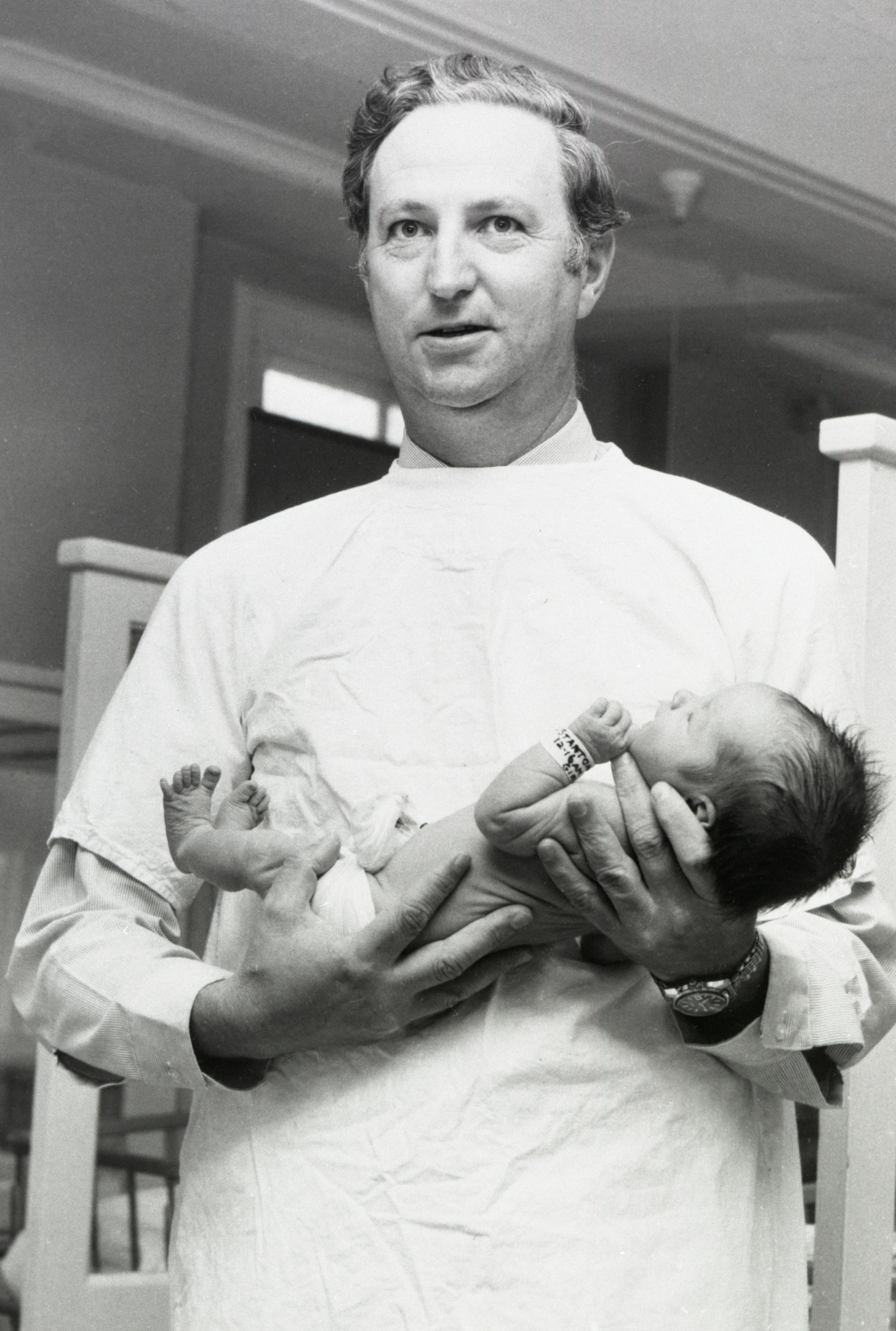 A man in a gown gently holds a newborn baby in a hospital setting, suggesting a caring moment possibly related to parenthood or medical care