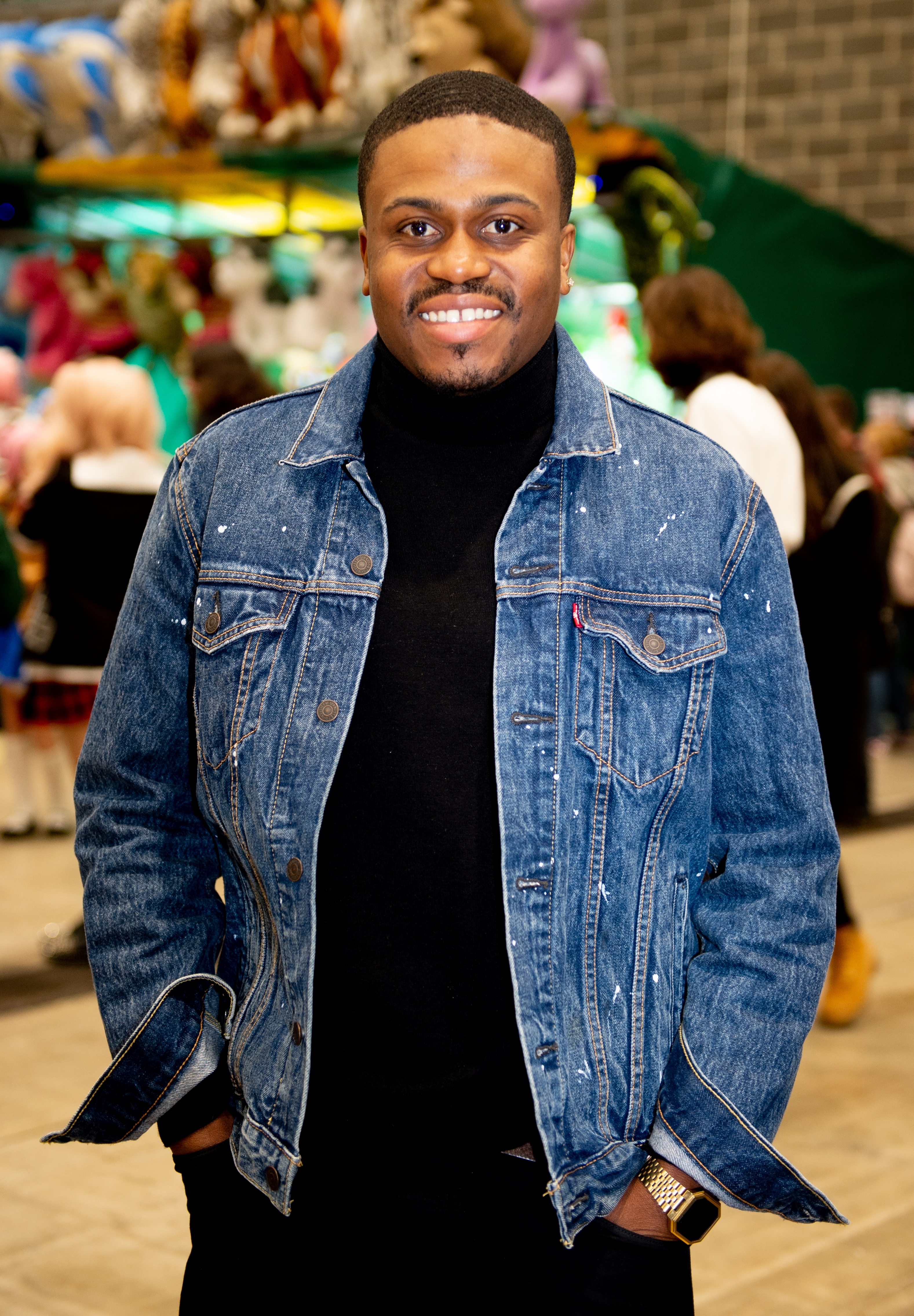 A person smiling, wearing a denim jacket over a black turtleneck, standing in a lively indoor setting with people in the background