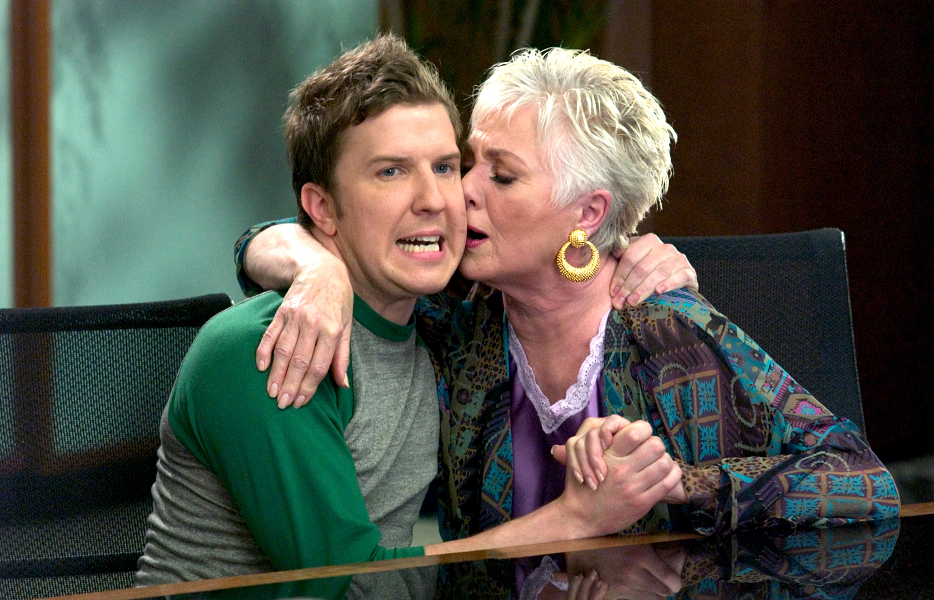 Older woman playfully hugging a younger man, both smiling. Woman wears a patterned jacket; man in casual attire. Playful, lighthearted interaction