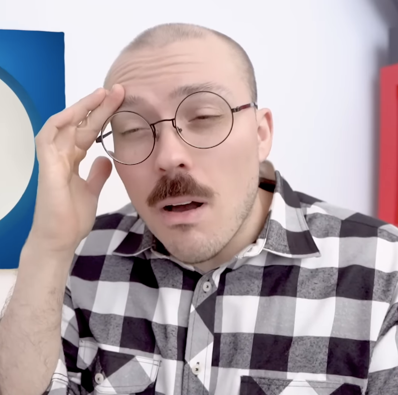 Man in a checkered shirt with glasses and a mustache, holding his head with a surprised or perplexed expression