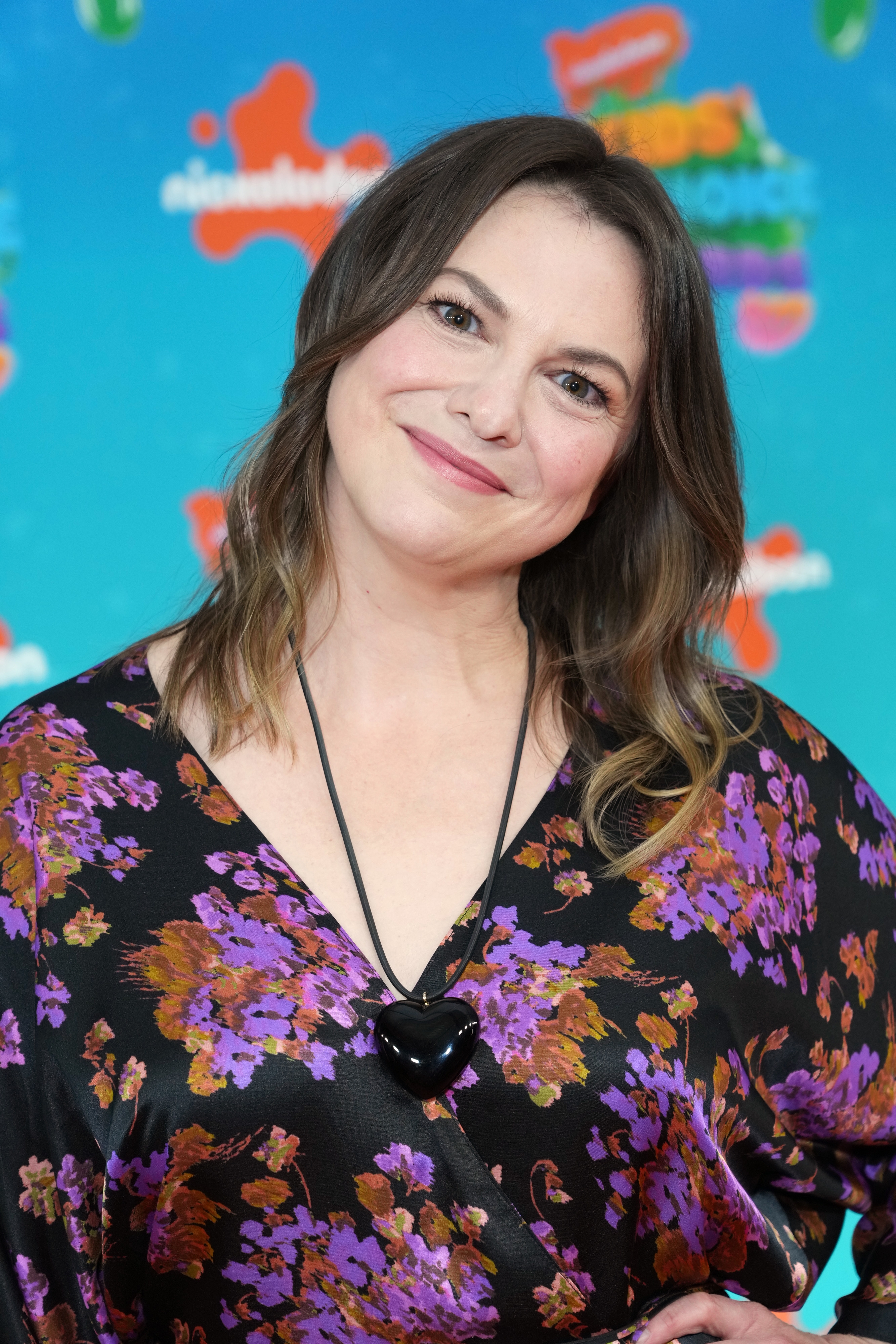 Person posing at an event, wearing a floral dress with a heart necklace, smiling against a branded backdrop