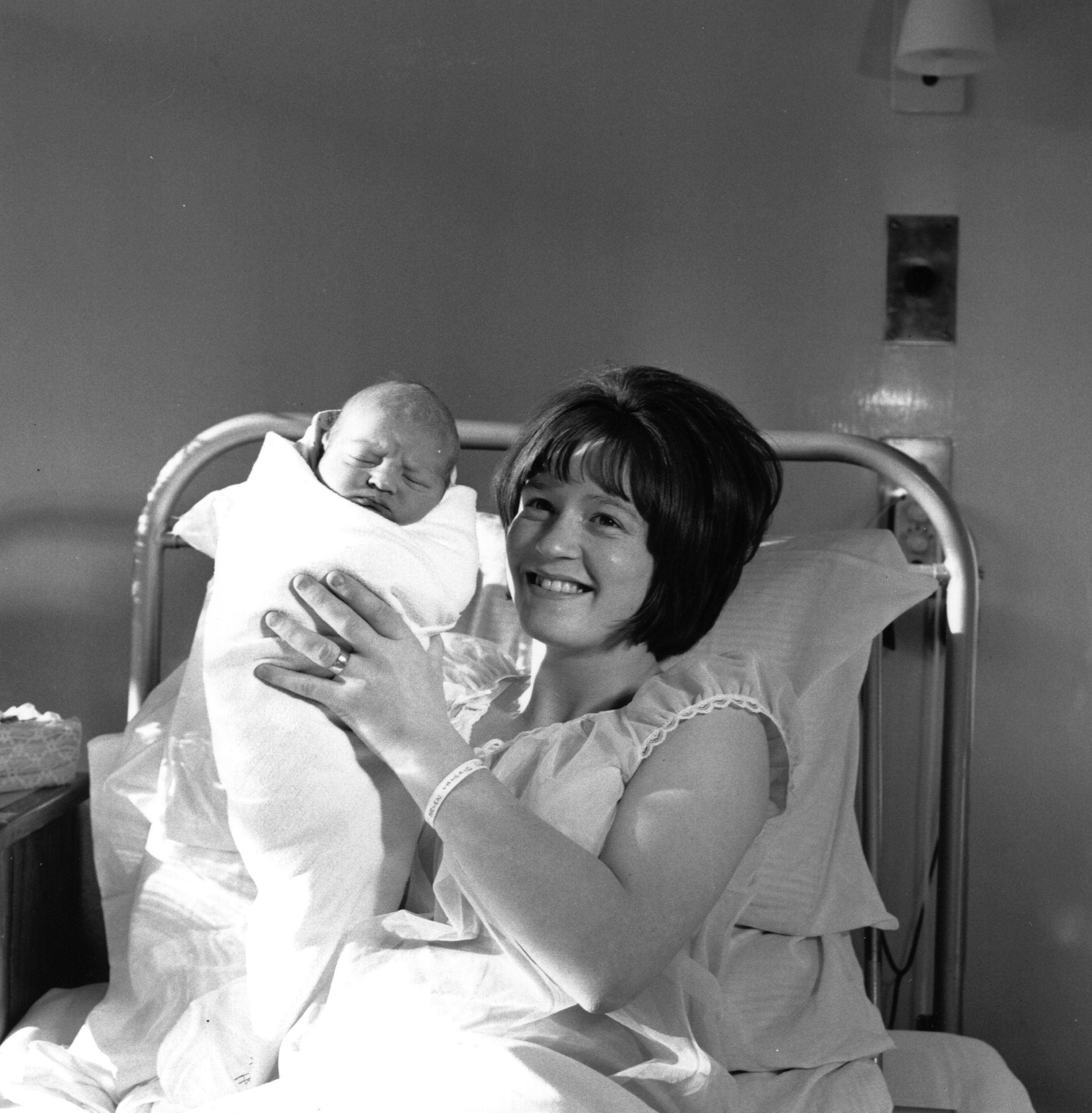 A mother joyfully holds her newborn in a hospital bed, both wrapped in blankets, symbolizing a moment of early parenthood