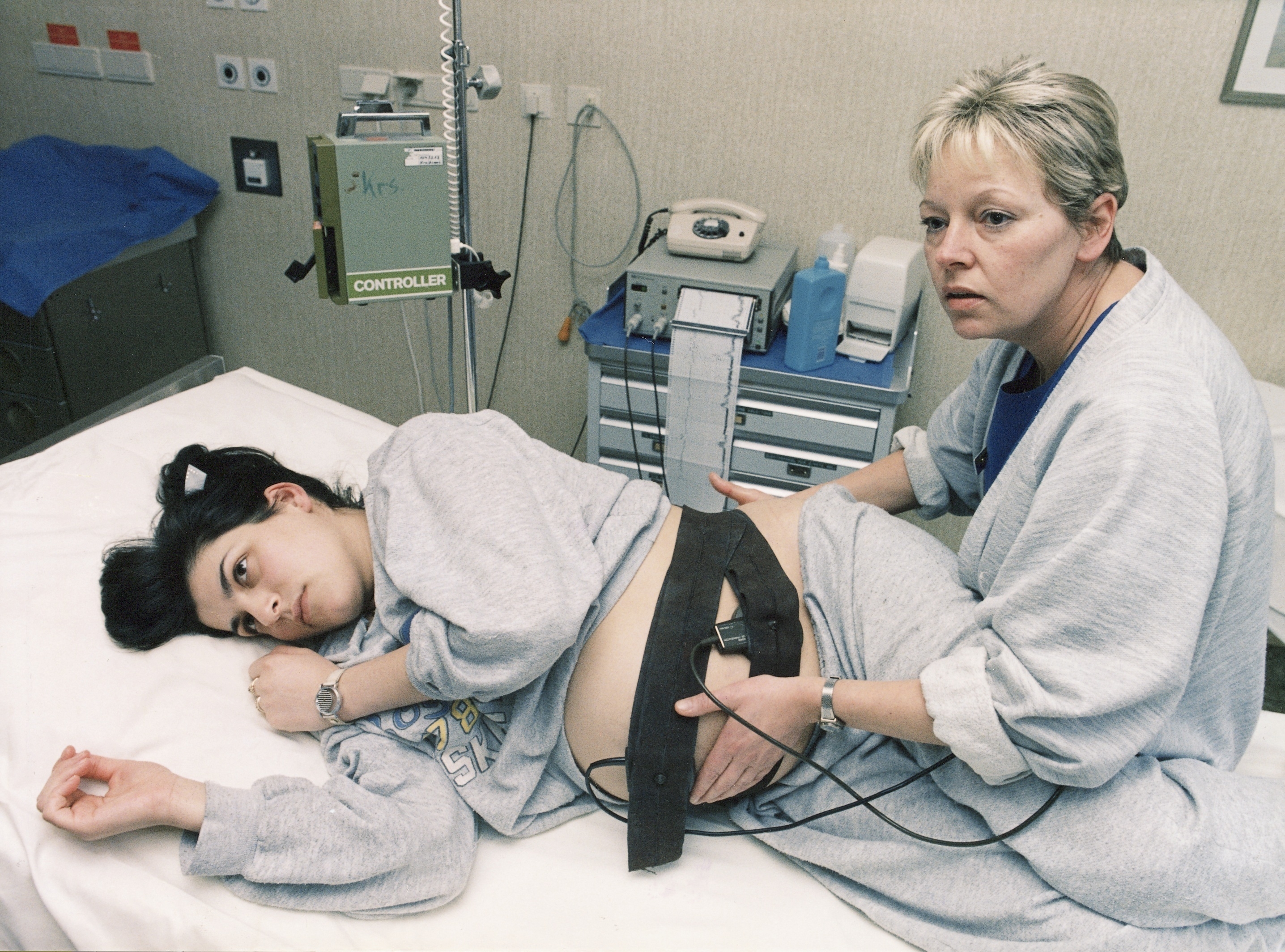A pregnant woman on a hospital bed undergoes monitoring with medical equipment, attended by a nurse