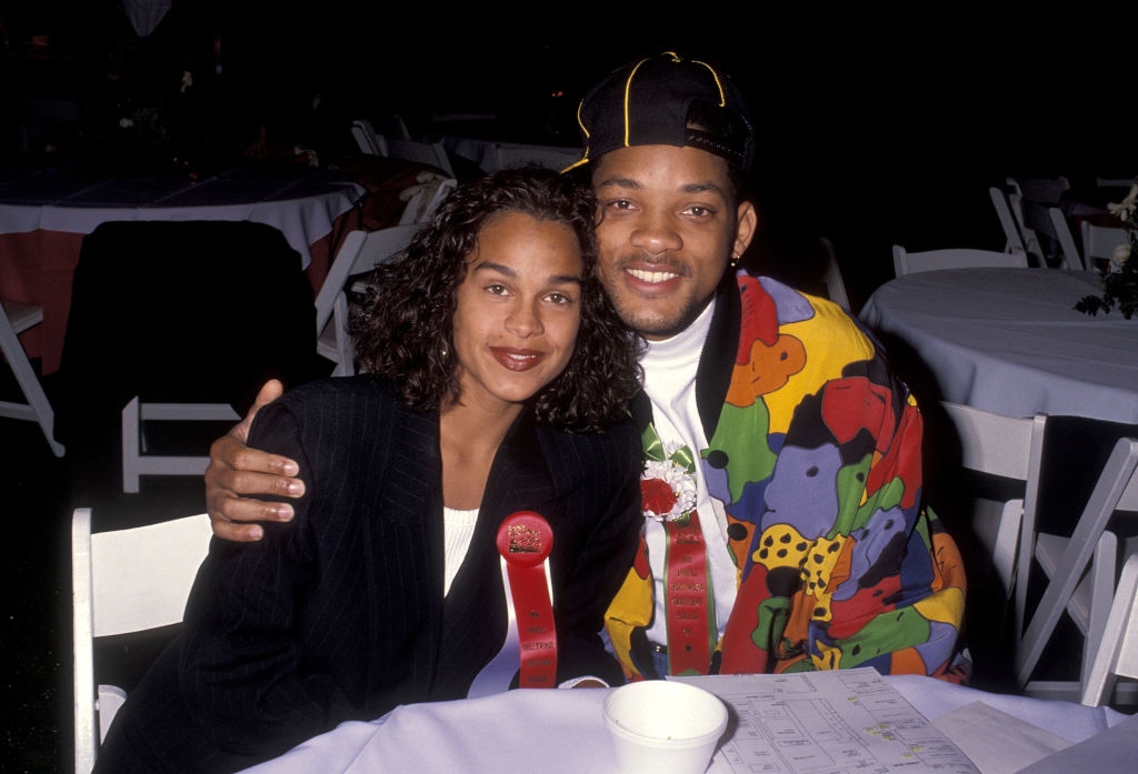 Two people seated at an event, smiling. One wears a colorful, patterned jacket and cap, the other wears a dark blazer. They both have award ribbons
