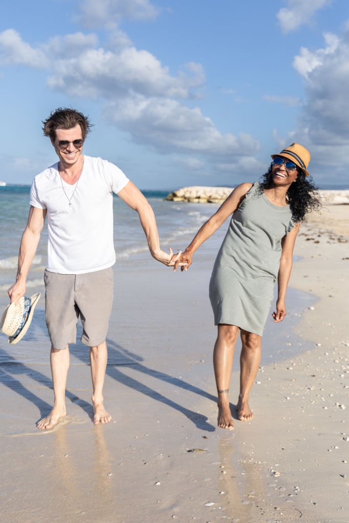 Two people smiling and holding hands while walking barefoot on a sandy beach, one wearing a casual dress and sunhat, and the other in a T-shirt and shorts
