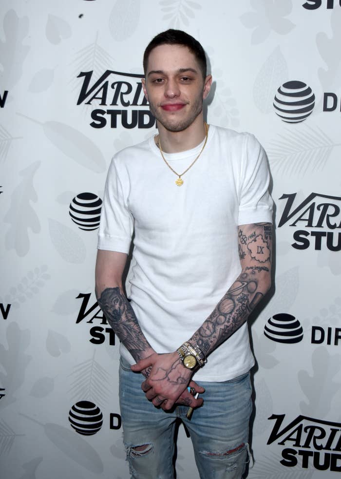 Person in a casual white t-shirt and jeans with visible tattoos on arms at a studio event backdrop