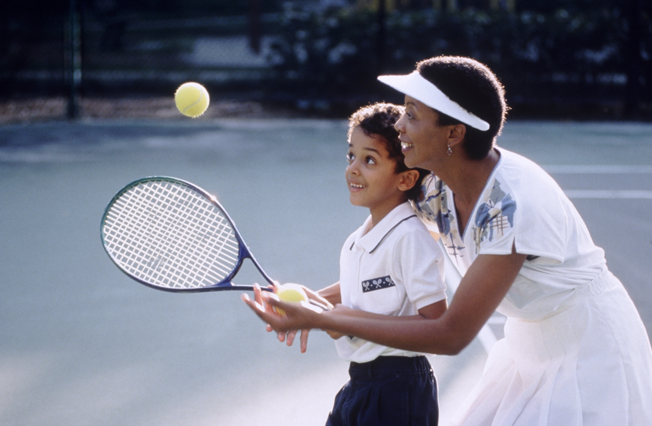 Woman guides child on tennis court, both holding a racket, smiling while practicing a swing