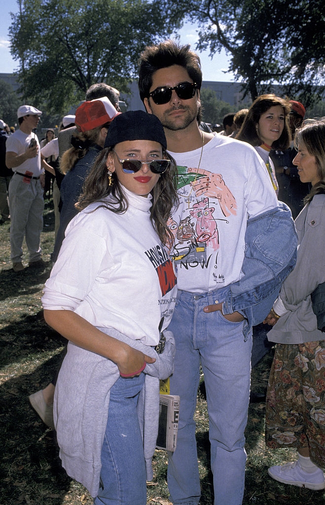 A casually dressed pair wearing graphic T-shirts and jeans poses outdoors at a celebrity event. They both wear sunglasses and appear relaxed