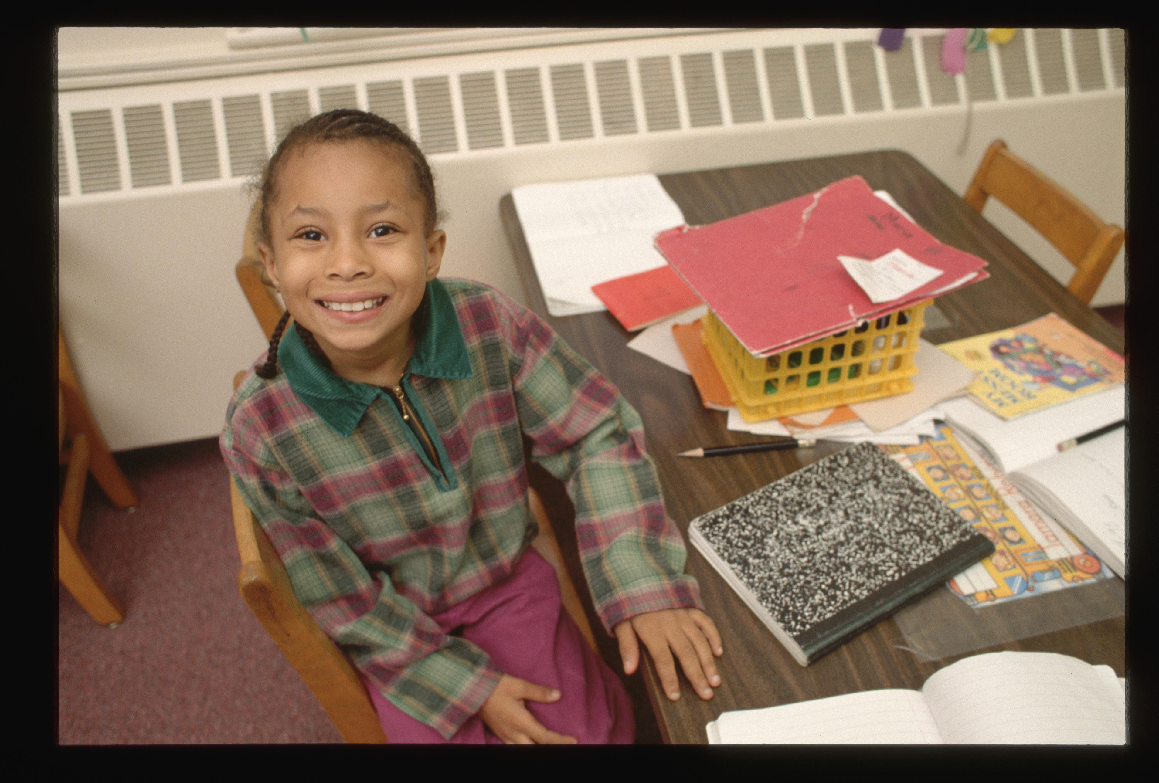 Child smiling at a classroom desk with open notebook and colorful papers