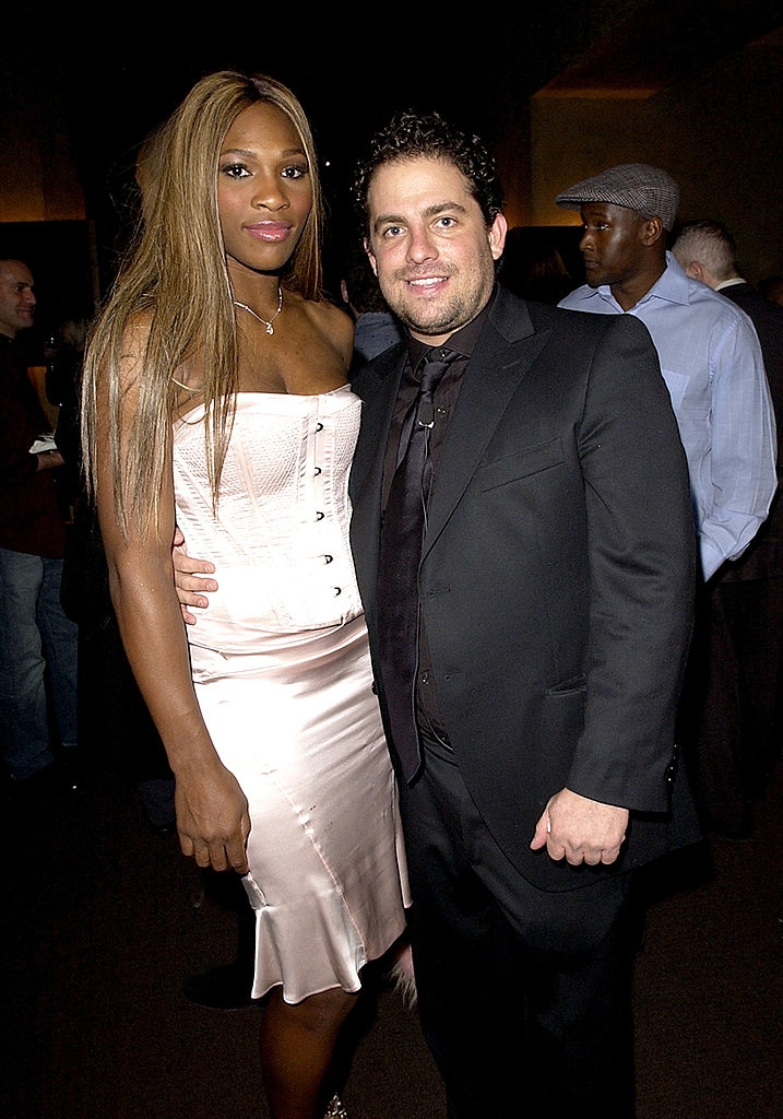 Two people pose together at an event. One wears a fitted corset top and satin skirt, while the other wears a dark suit