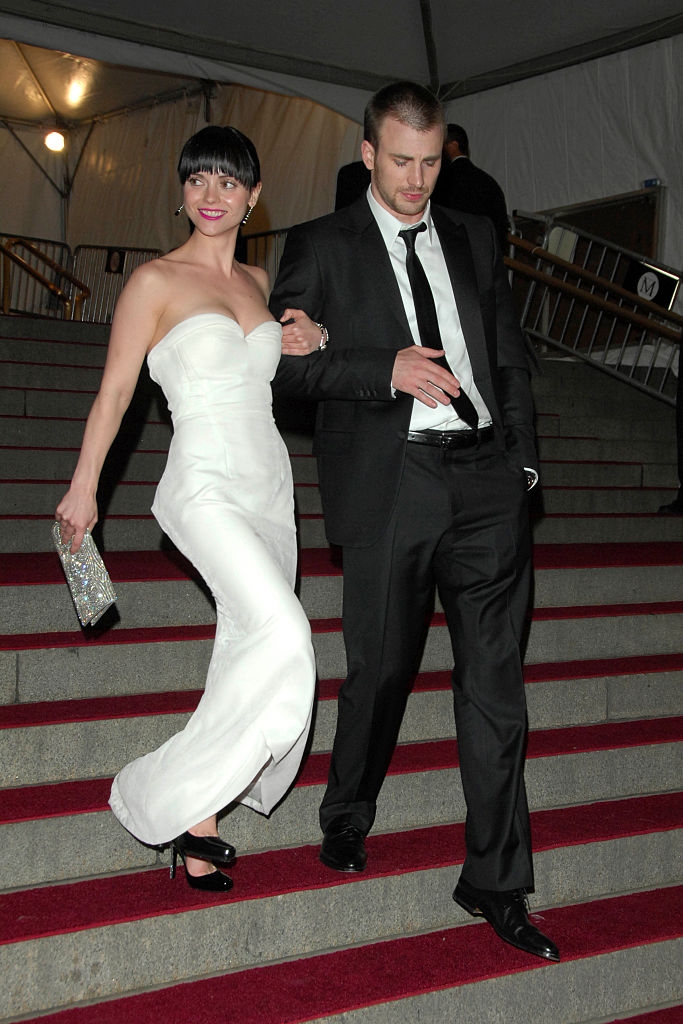 A woman in a strapless gown and a man in a suit walk arm-in-arm down a red carpeted staircase, both smiling and holding evening accessories