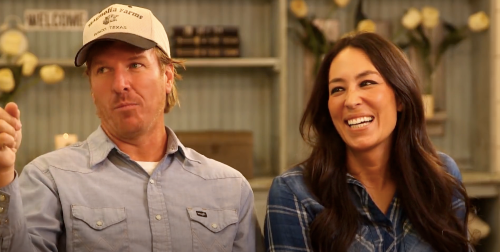 Two people sitting and smiling, one wears a baseball cap and casual shirt, the other has long hair and a plaid shirt, in a cozy room setting