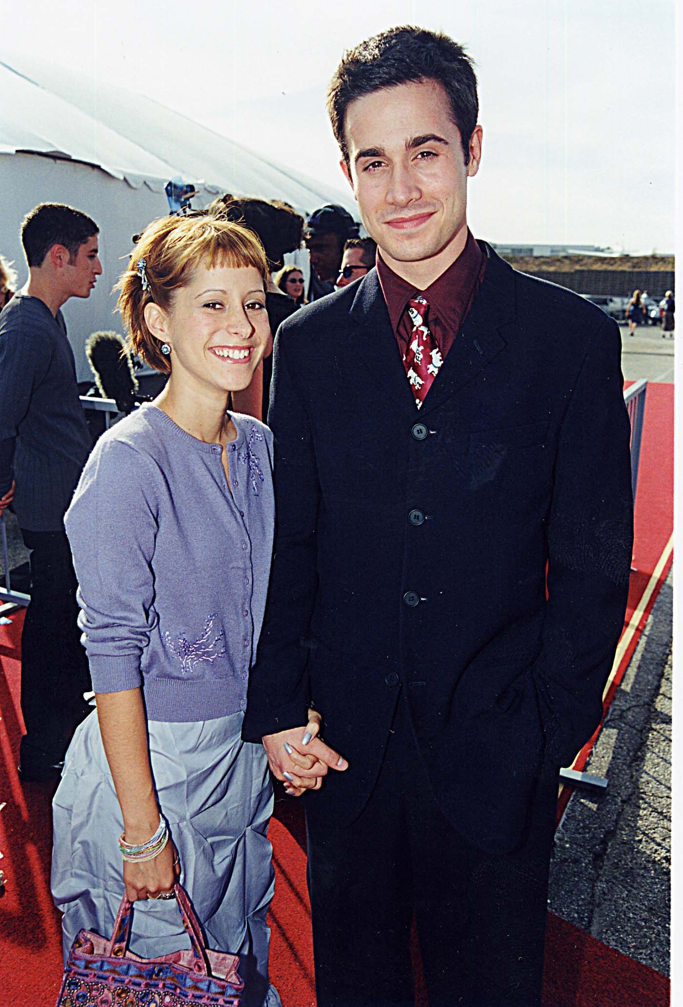 Two people pose on a red carpet. One wears a tailored dark suit with a patterned tie, and the other is in a light outfit with a ruffled skirt