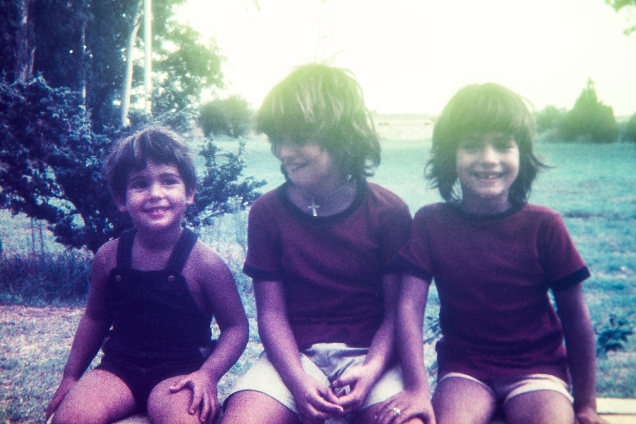 Three young children sit outdoors on a bench, smiling. They are wearing casual summer clothing. Trees and grass are visible in the background