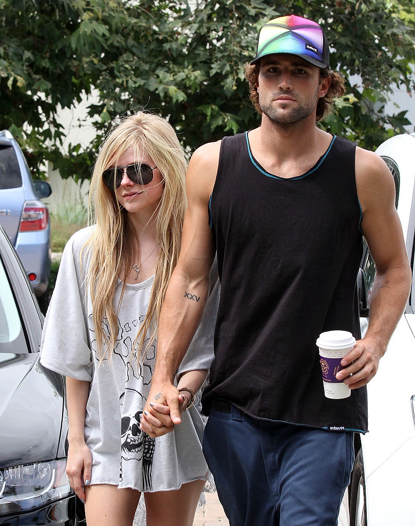 Celebrity couple holding hands outdoors; she wears a loose graphic tee, and he wears a tank top with a colorful cap, holding a coffee cup