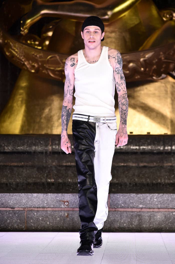 Person with tattoos in a white tank top and two-tone black and white pants walking a runway