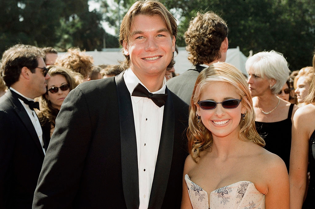 A man in a tuxedo and a woman in a strapless gown with a floral pattern smile at a formal outdoor event