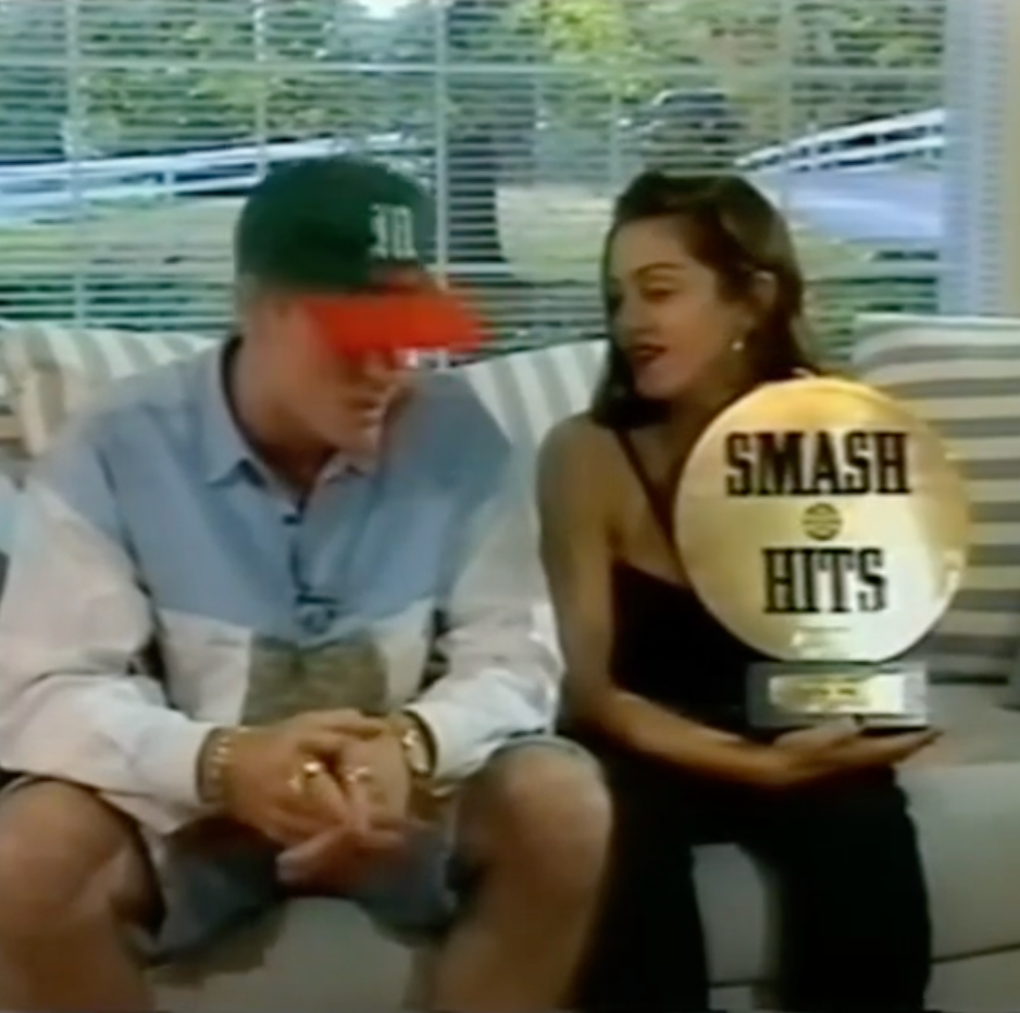 Two individuals sit together on a couch, one holding a "Smash Hits" award trophy. The person on the right wears a sleeveless top