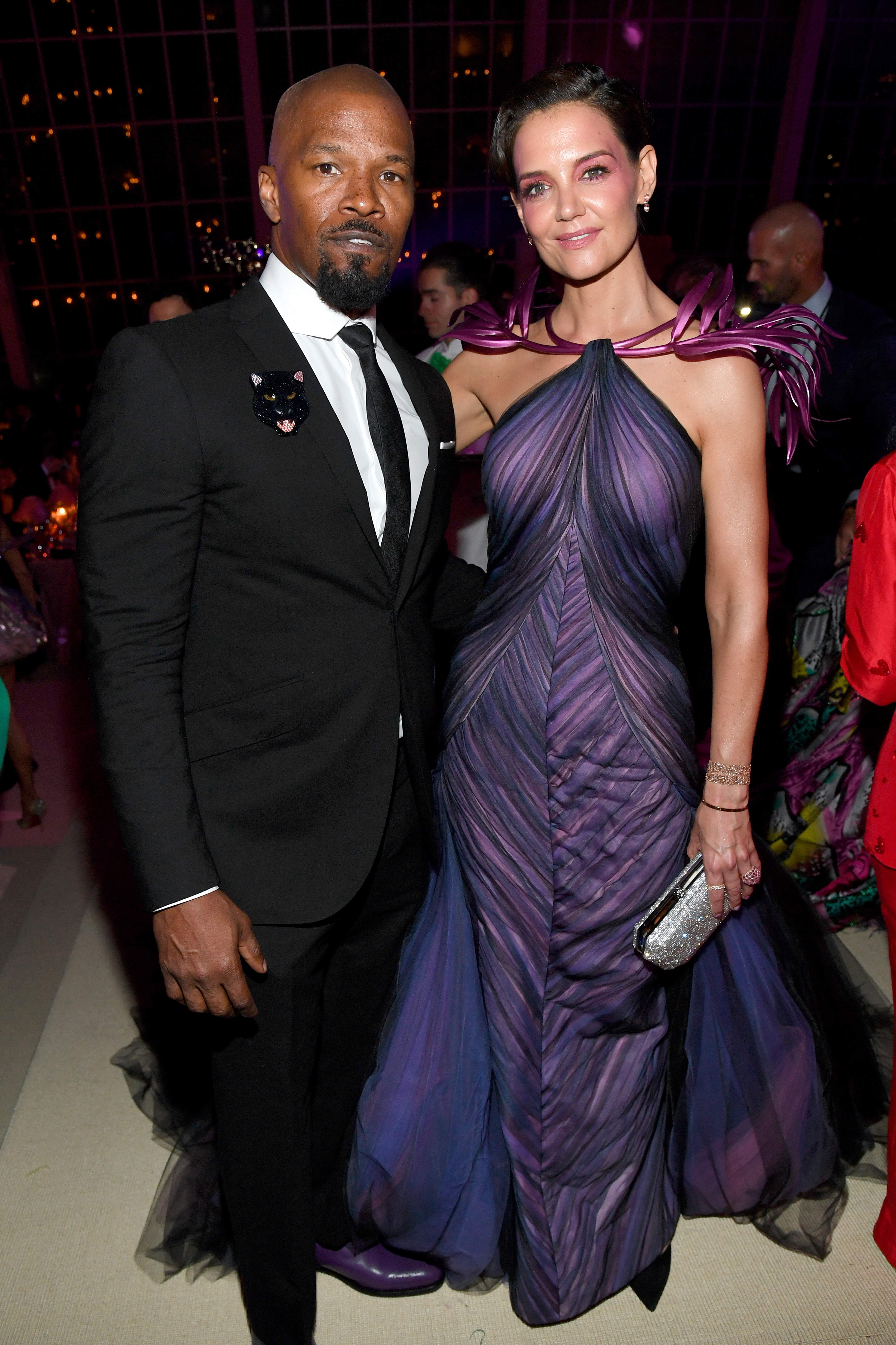 Two celebrities pose on the red carpet; one in a dark suit, the other in a purple gown with feathered shoulders, holding a clutch