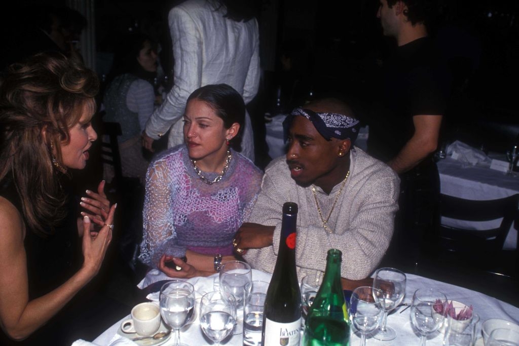 Three people sit at a table engaged in conversation. One person wears a lace top; another a bandana and patterned jacket. Wine and water are on the table