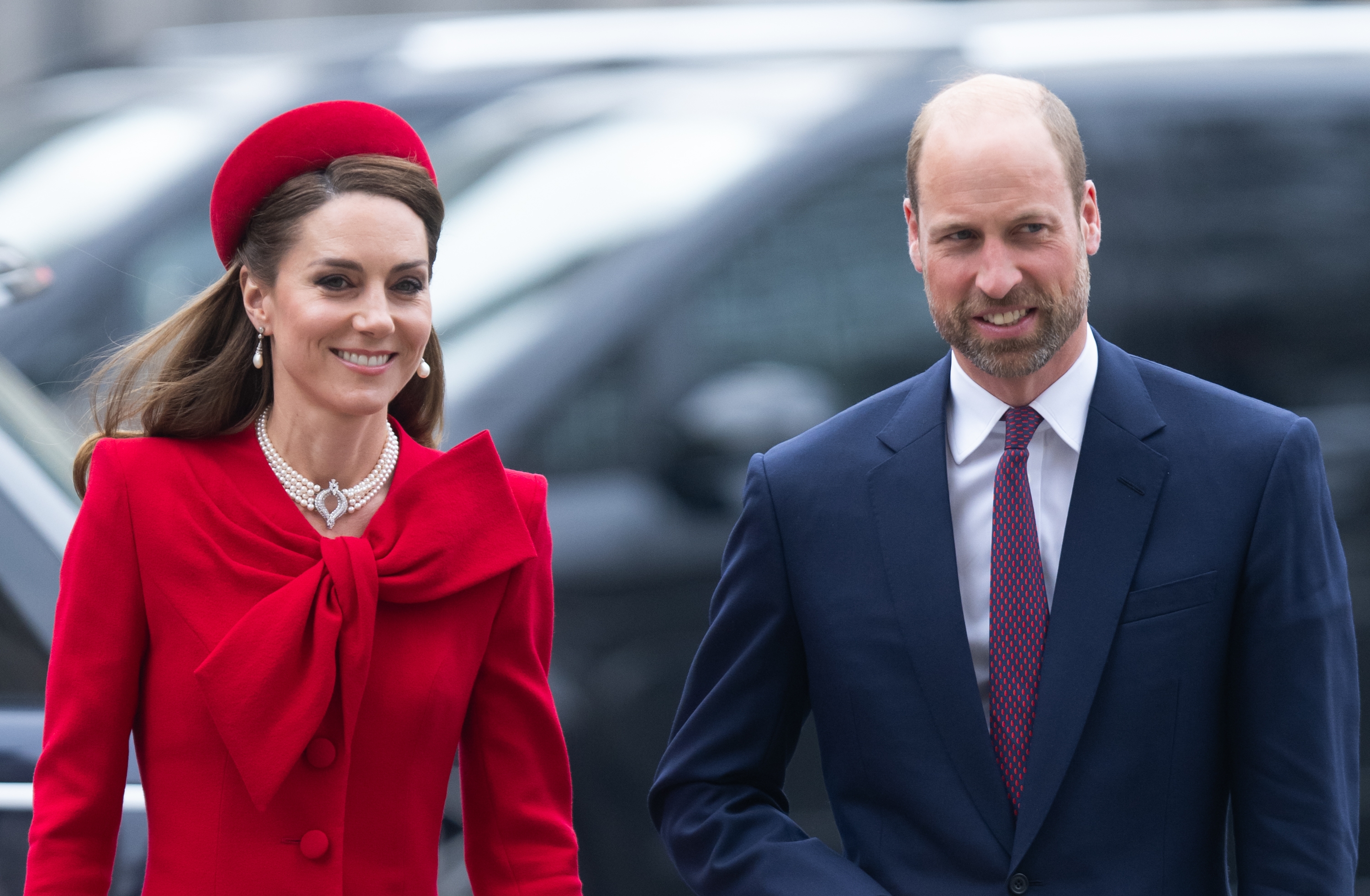A woman wearing a formal red outfit and a man in a blue suit walk side by side, smiling, in an outdoor setting