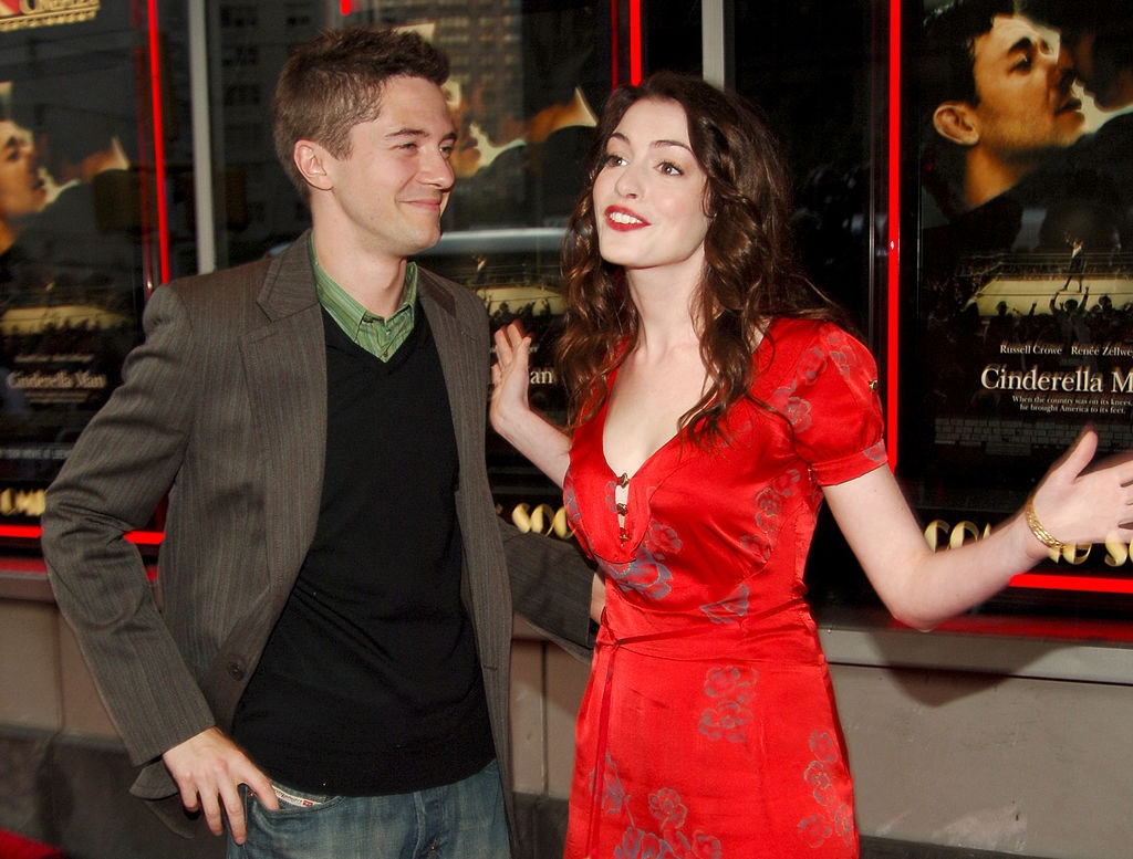 Two people on a red carpet; the woman wears a floral dress, and the man is in a blazer with a casual shirt. Movie posters in the background