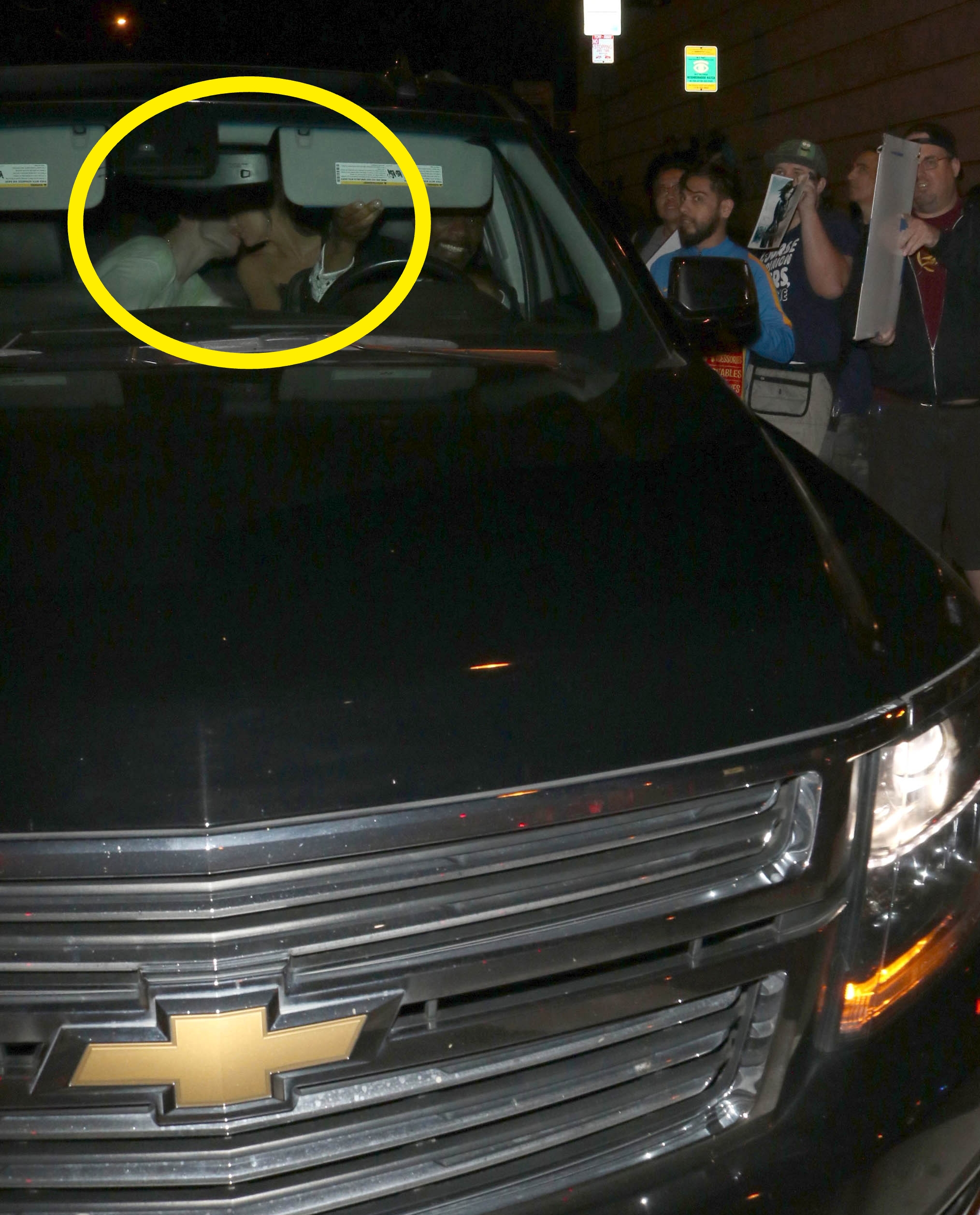 A celebrity is seen inside a parked SUV surrounded by paparazzi and fans holding cameras and signs on a dimly lit street