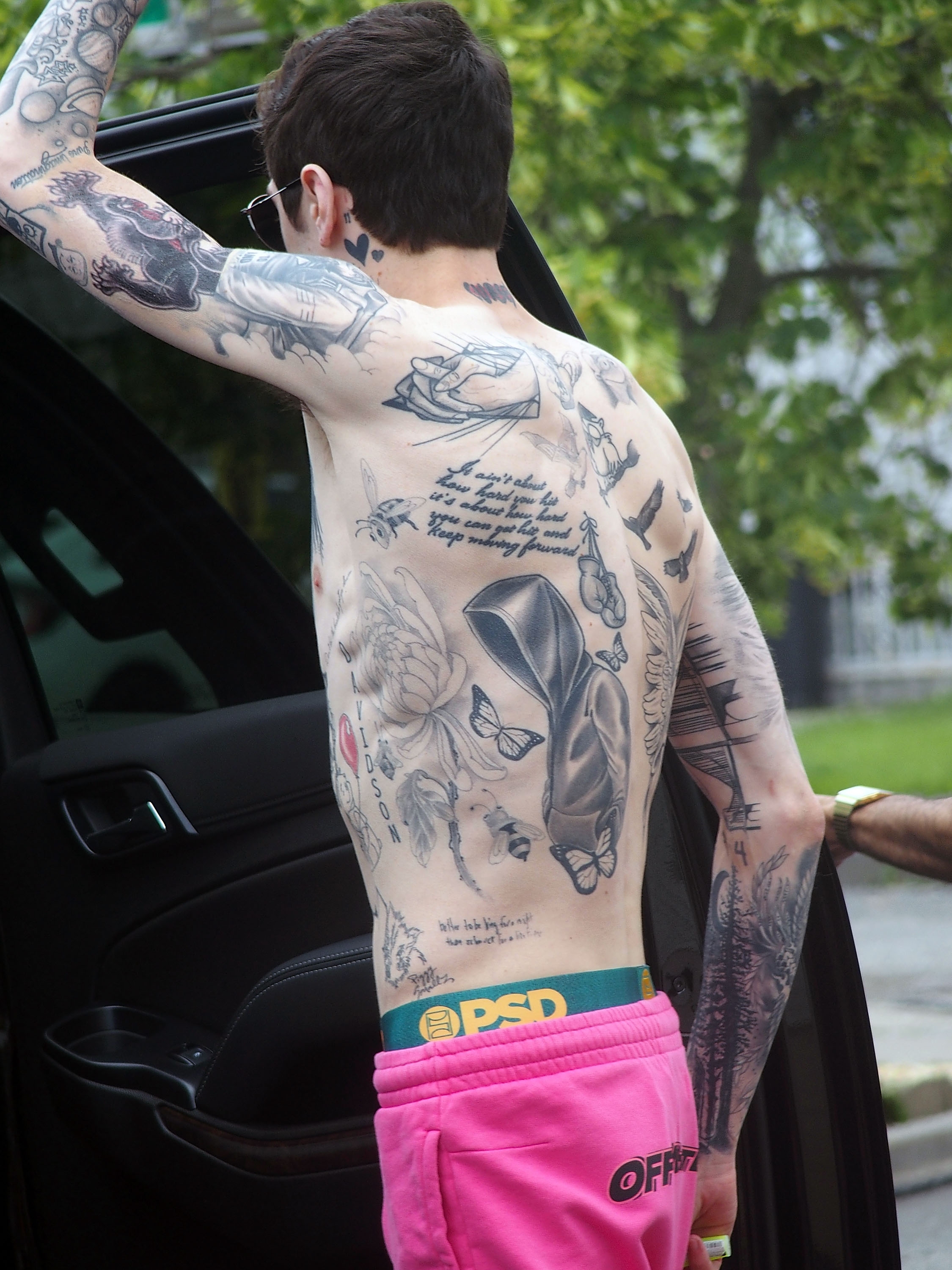 A person with extensive tattoos on their back and arms is stepping out of a vehicle, wearing pink pants and sunglasses outdoors