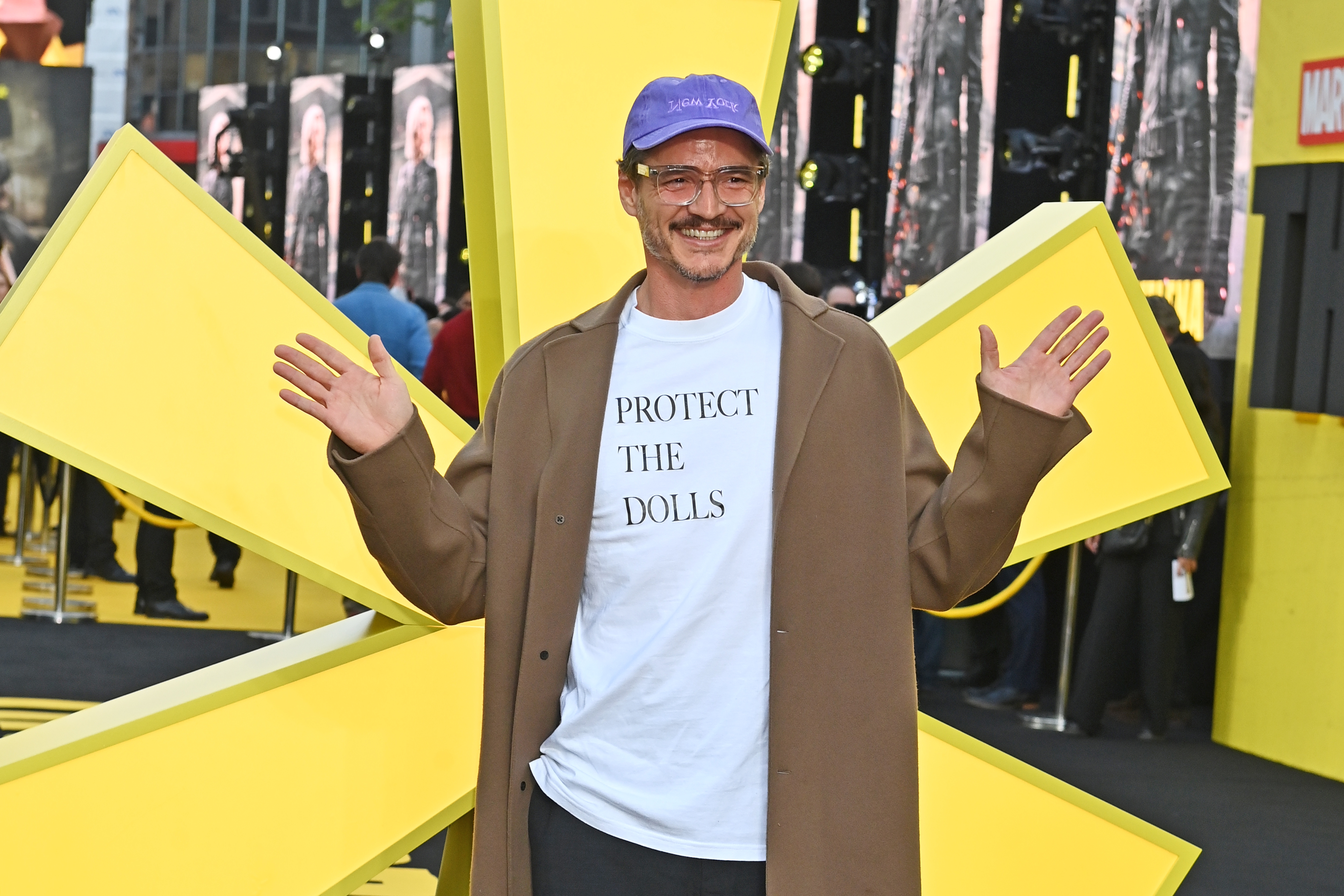 Celebrity on a yellow star-shaped carpet, wearing a casual outfit with a light coat, printed T-shirt, and cap, posing with hands up