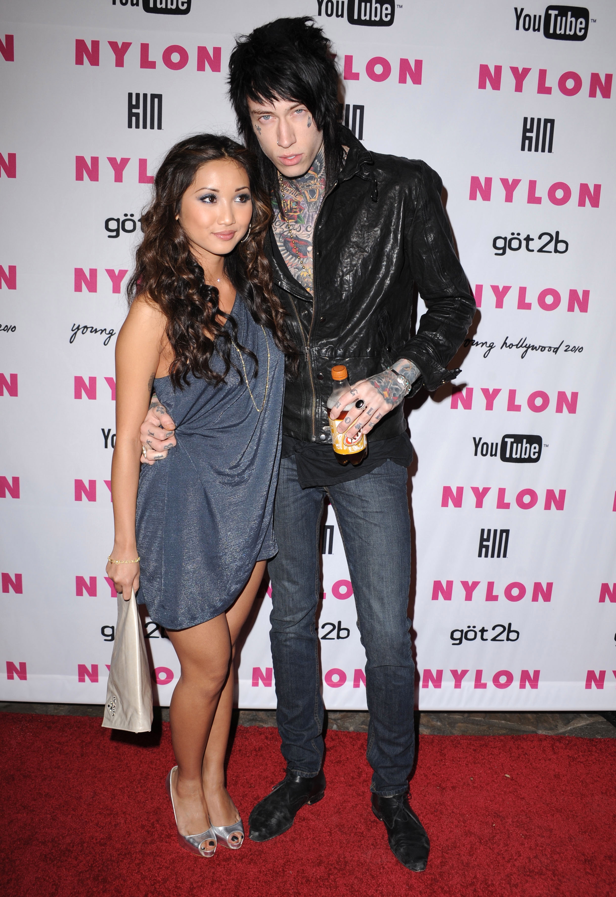 Two people pose on a red carpet. The person on the left wears a short, draped dress; the person on the right wears a leather jacket with tattoos visible