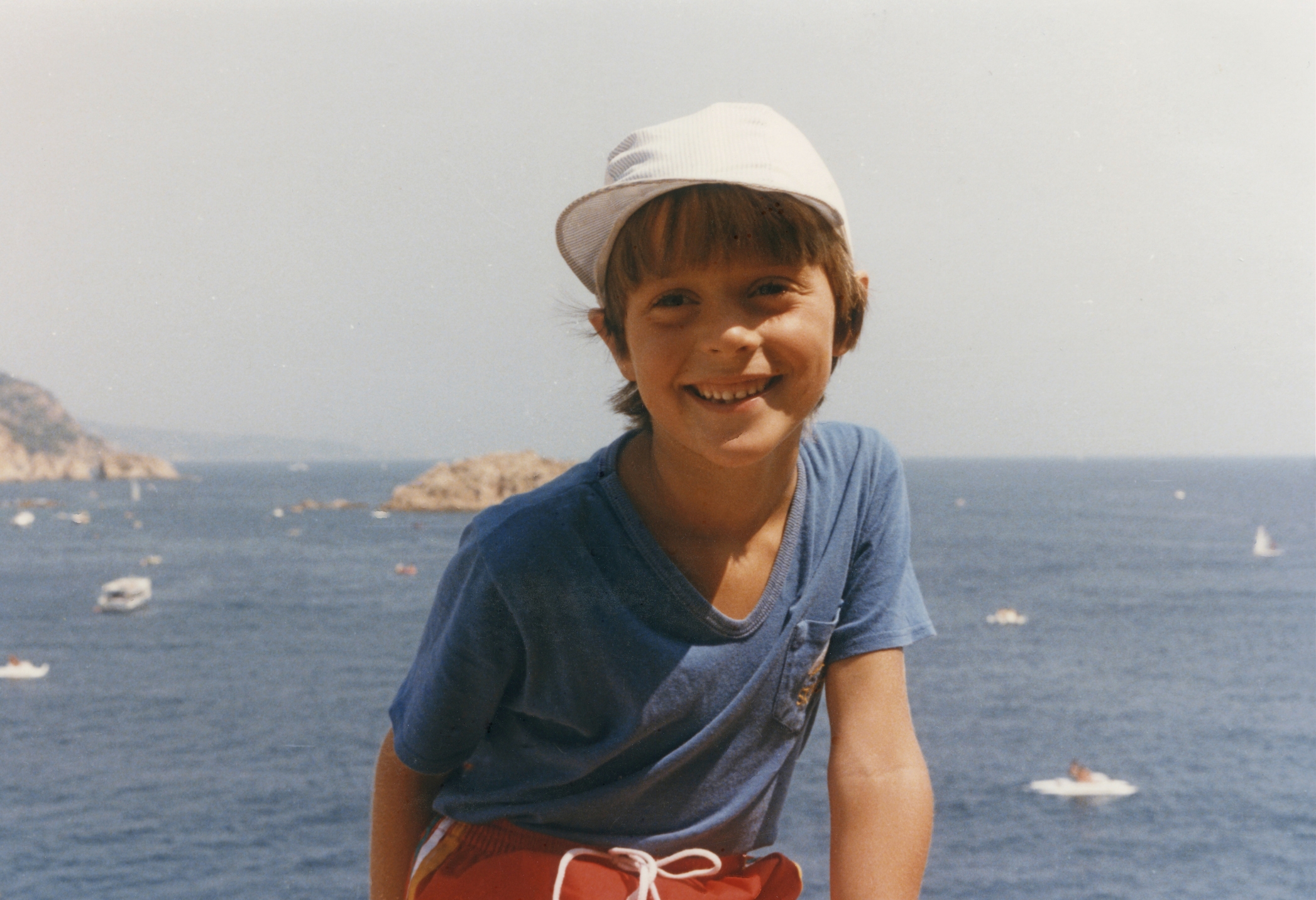 Child smiling by the sea, wearing a light cap and a casual shirt, with boats in the background