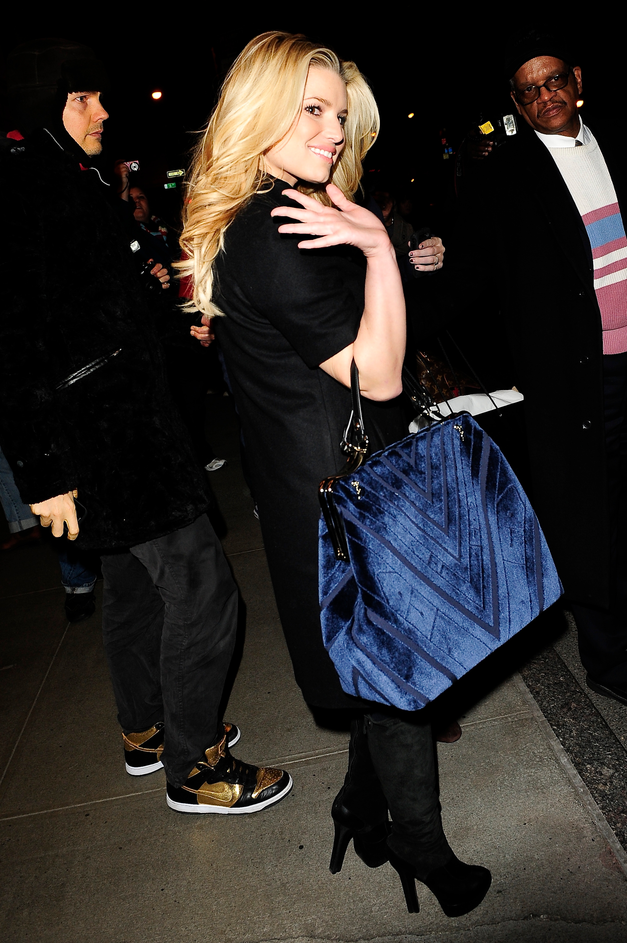 A woman on the street poses for photos, wearing a black coat, holding a large patterned bag, and smiling over her shoulder