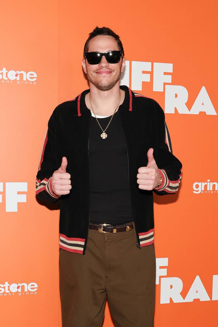 Person in casual attire with a jacket and sunglasses, giving thumbs-up on a red carpet