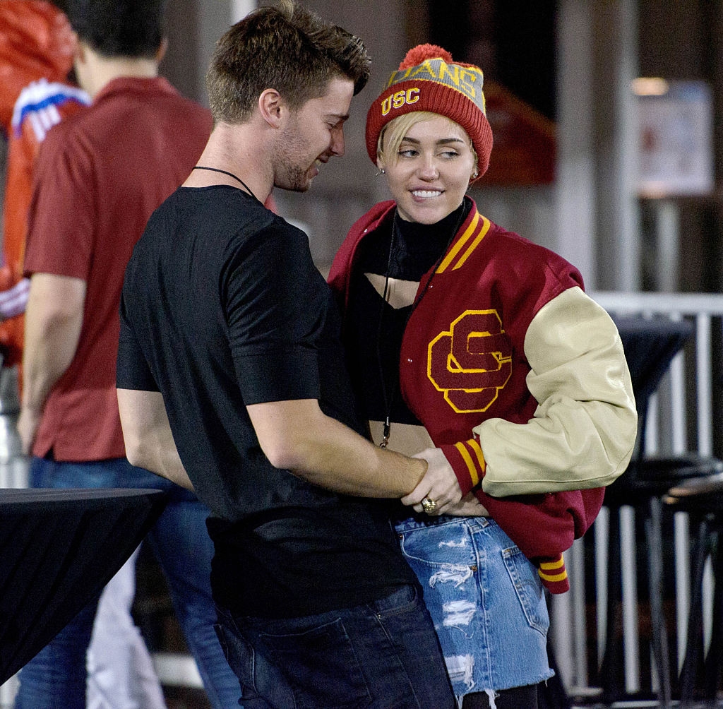 Two people stand close, smiling at each other. One wears a USC jacket and beanie with ripped jeans, while the other wears a black top and jeans