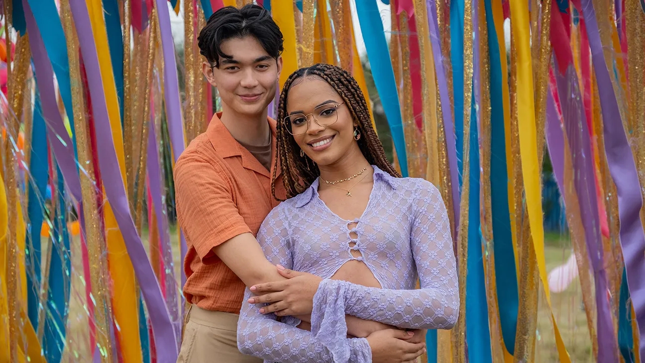 Dos personas posan abrazadas frente a un fondo de cintas coloridas. La persona de la derecha lleva una blusa de encaje