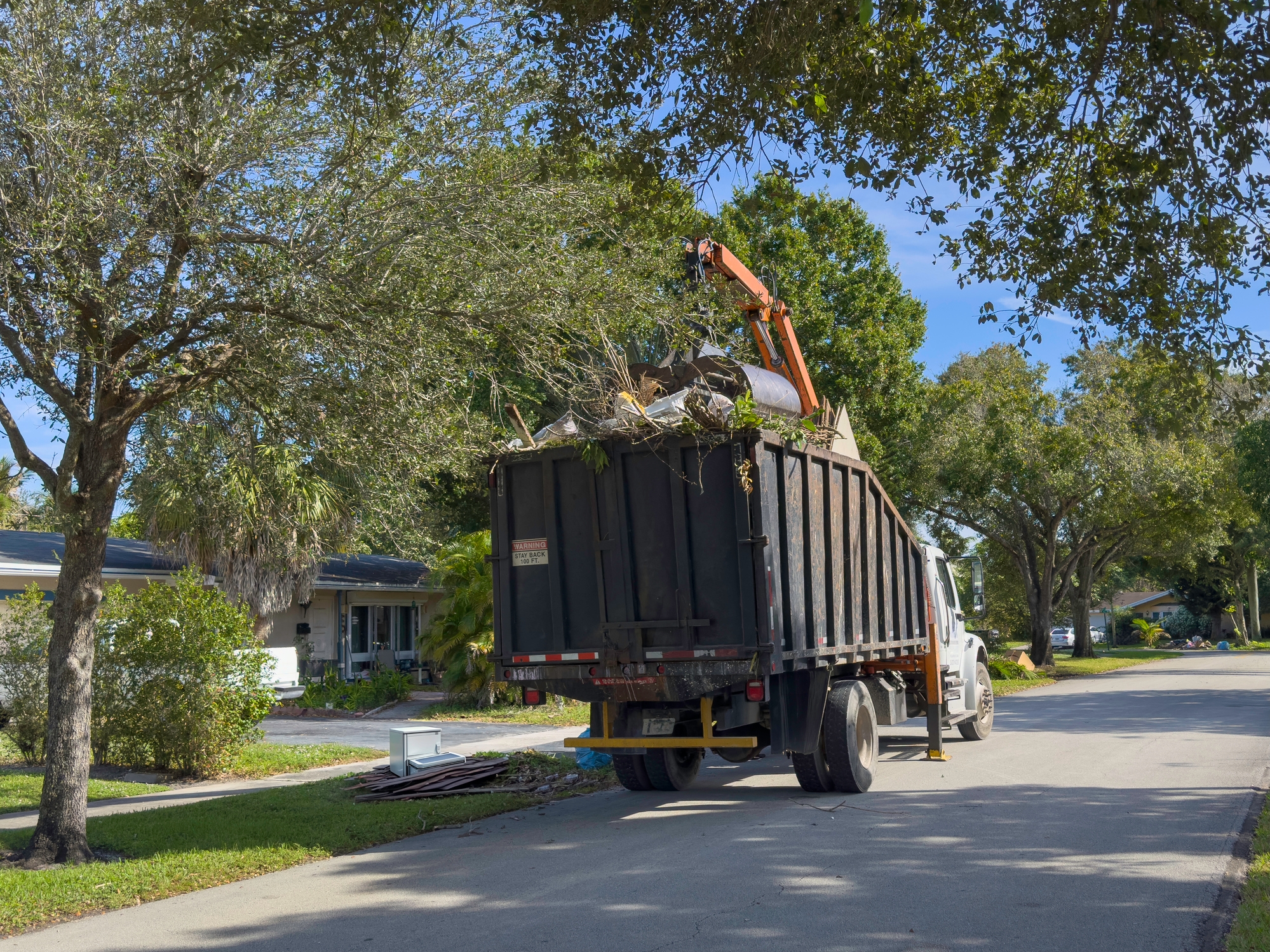 A truck mounted with a claw crane collects tree branches from a suburban street lined with houses and trees