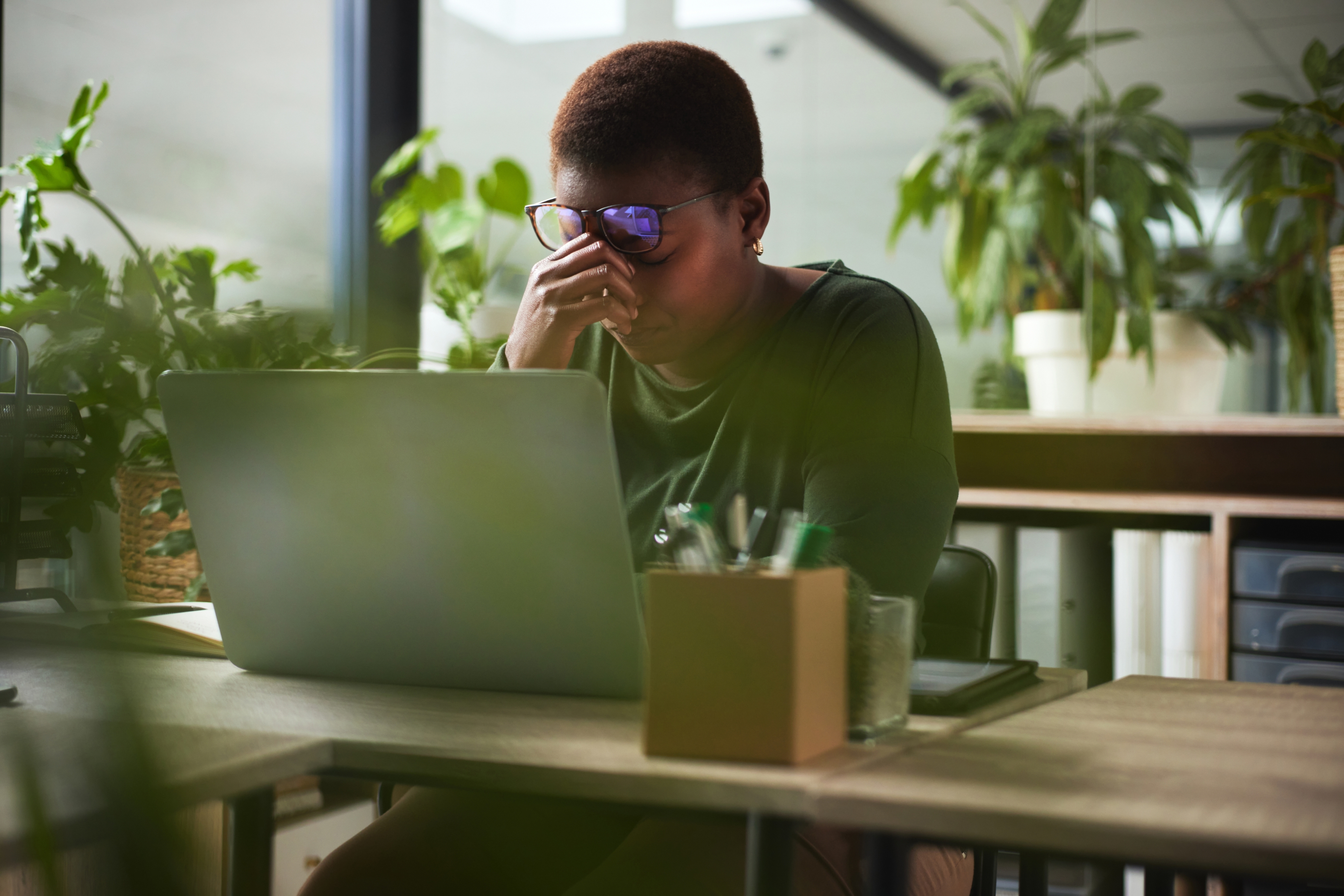Person sitting at a desk with a laptop, appearing stressed, holding their face with one hand in a plant-filled office space