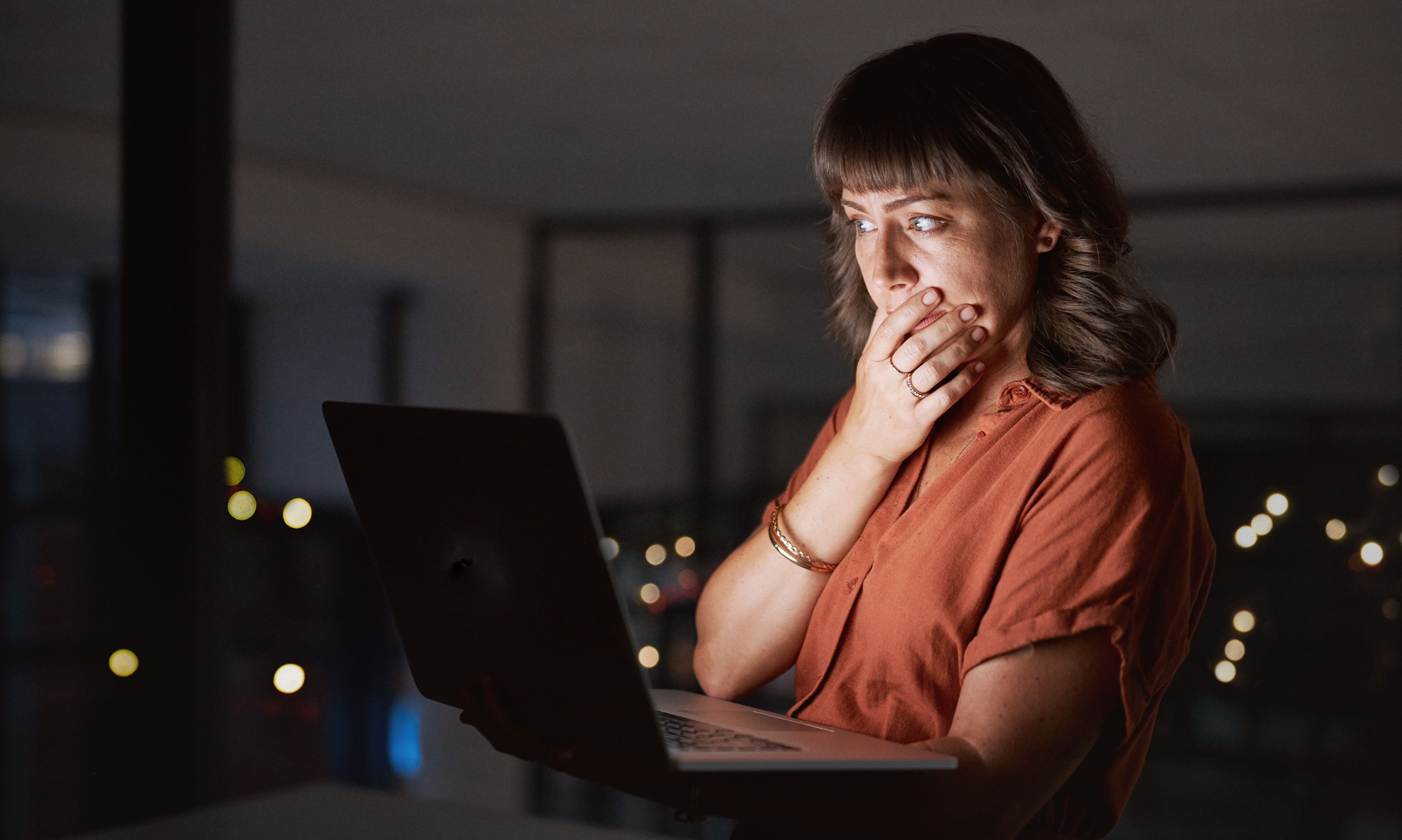 Person with a thoughtful expression, holding a laptop in dim light, implying deep contemplation or surprise in a private setting