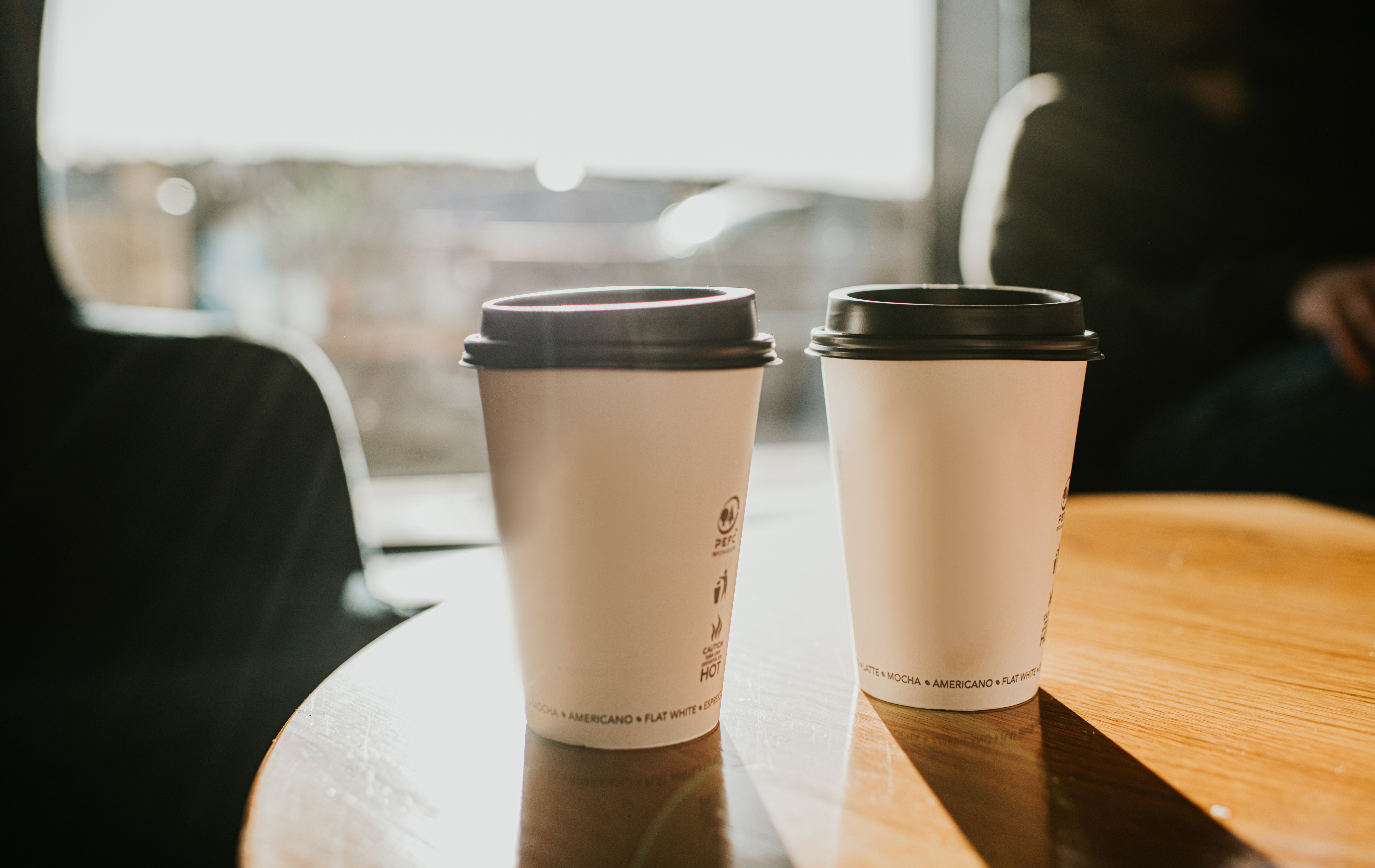 Two takeaway coffee cups on a wooden table in a sunlit room, suggesting a cozy setting for a romantic or intimate conversation